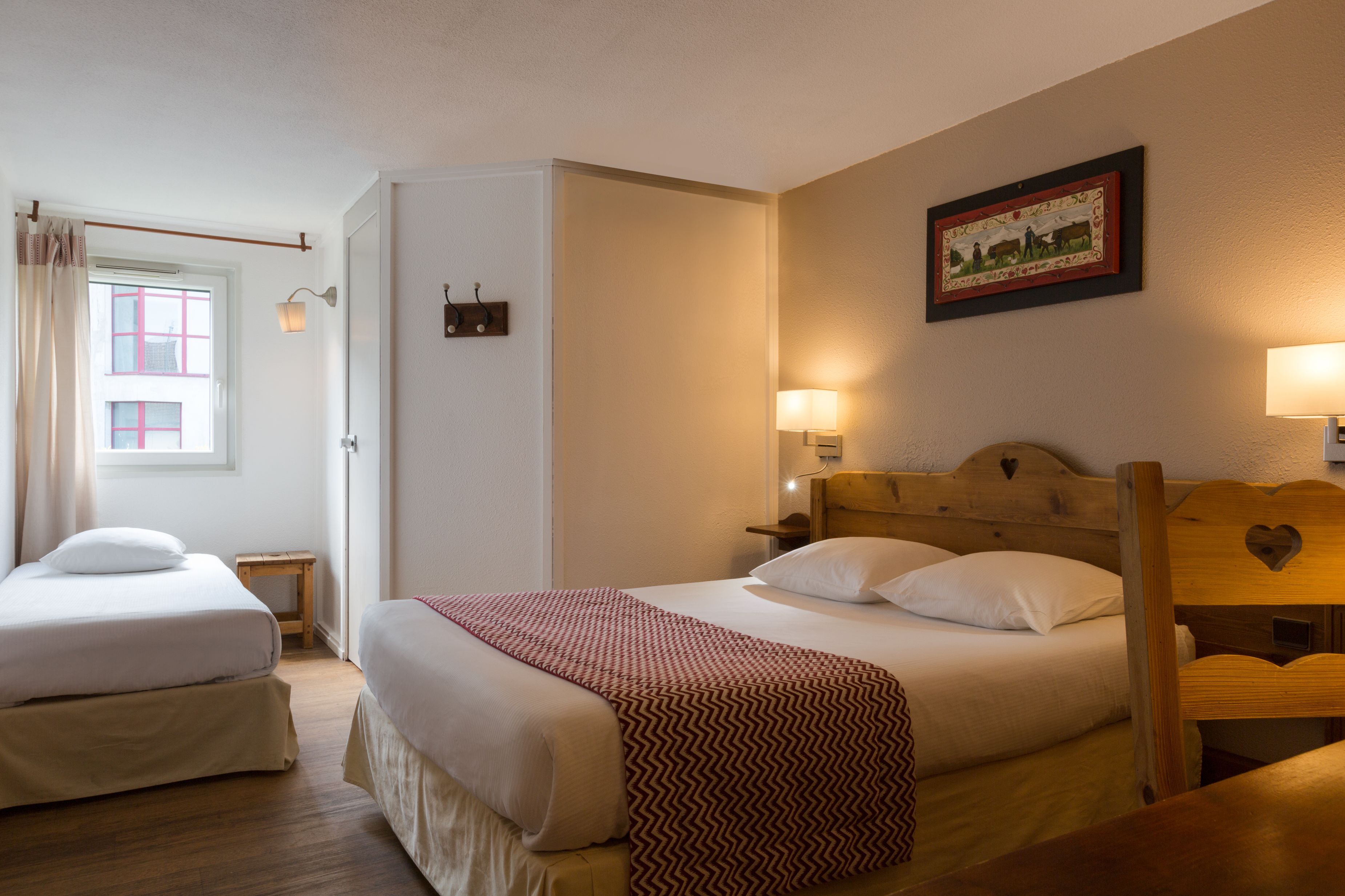 A room with two beds, a wooden headboard, a small table, a wall-mounted lamp, and a framed picture.