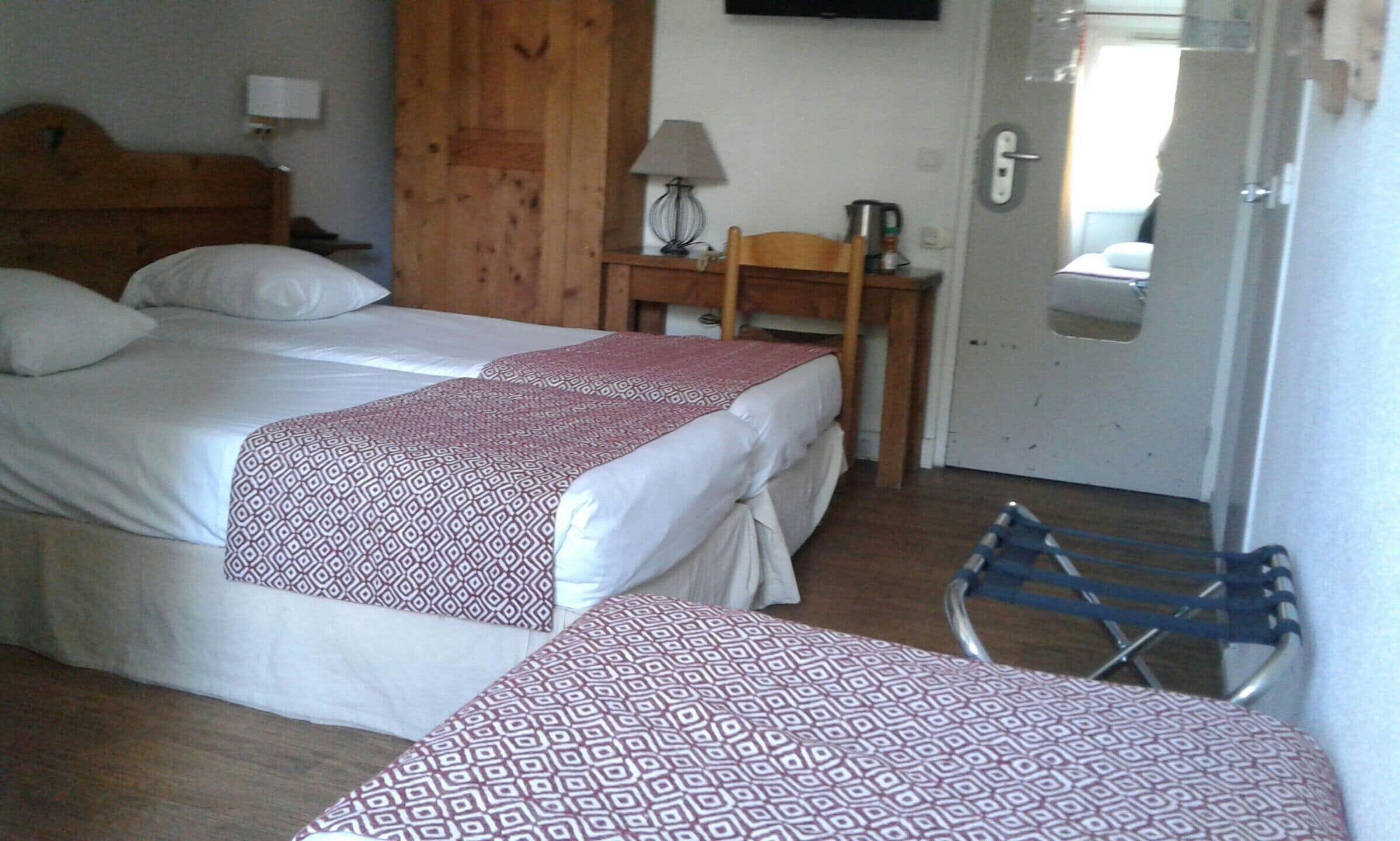 Ein Hotelzimmer mit zwei Betten, einem hölzernen Kopfteil, einem kleinen Tisch mit Lampe, einem Stuhl und einer Badezimmertür.