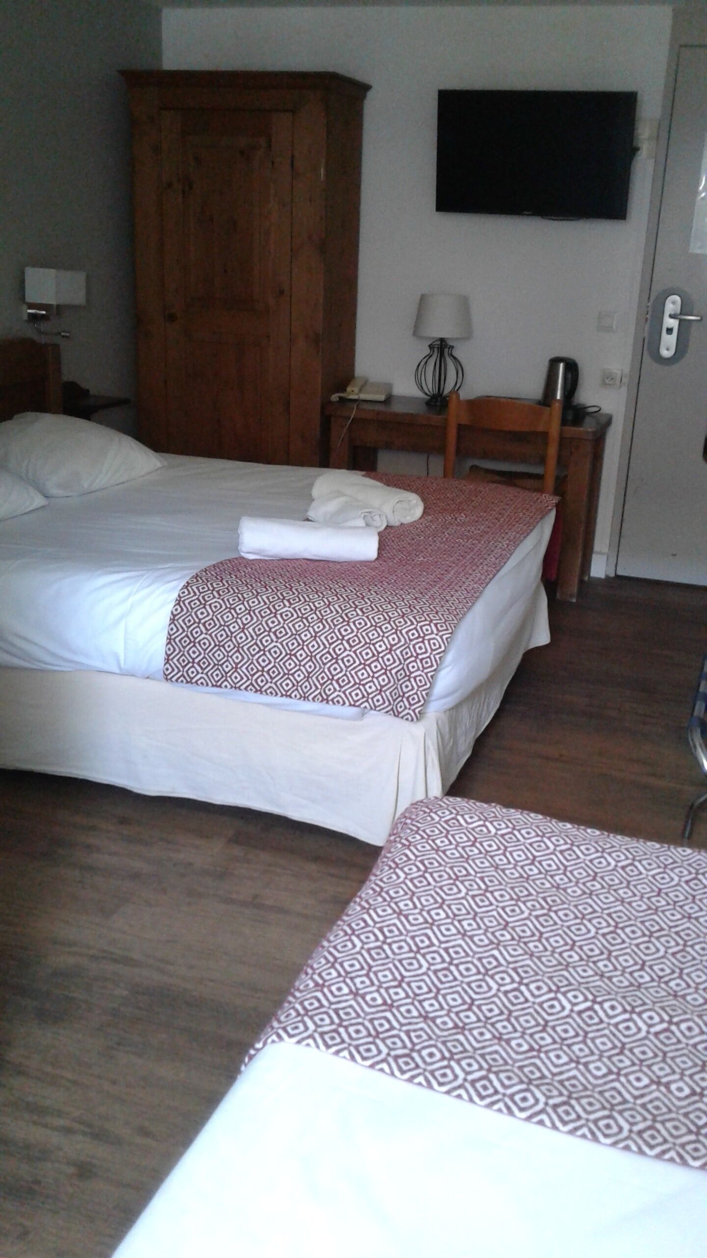 Une chambre d’hôtel avec deux lits, une armoire en bois, un bureau avec une lampe et un téléviseur fixé au mur.