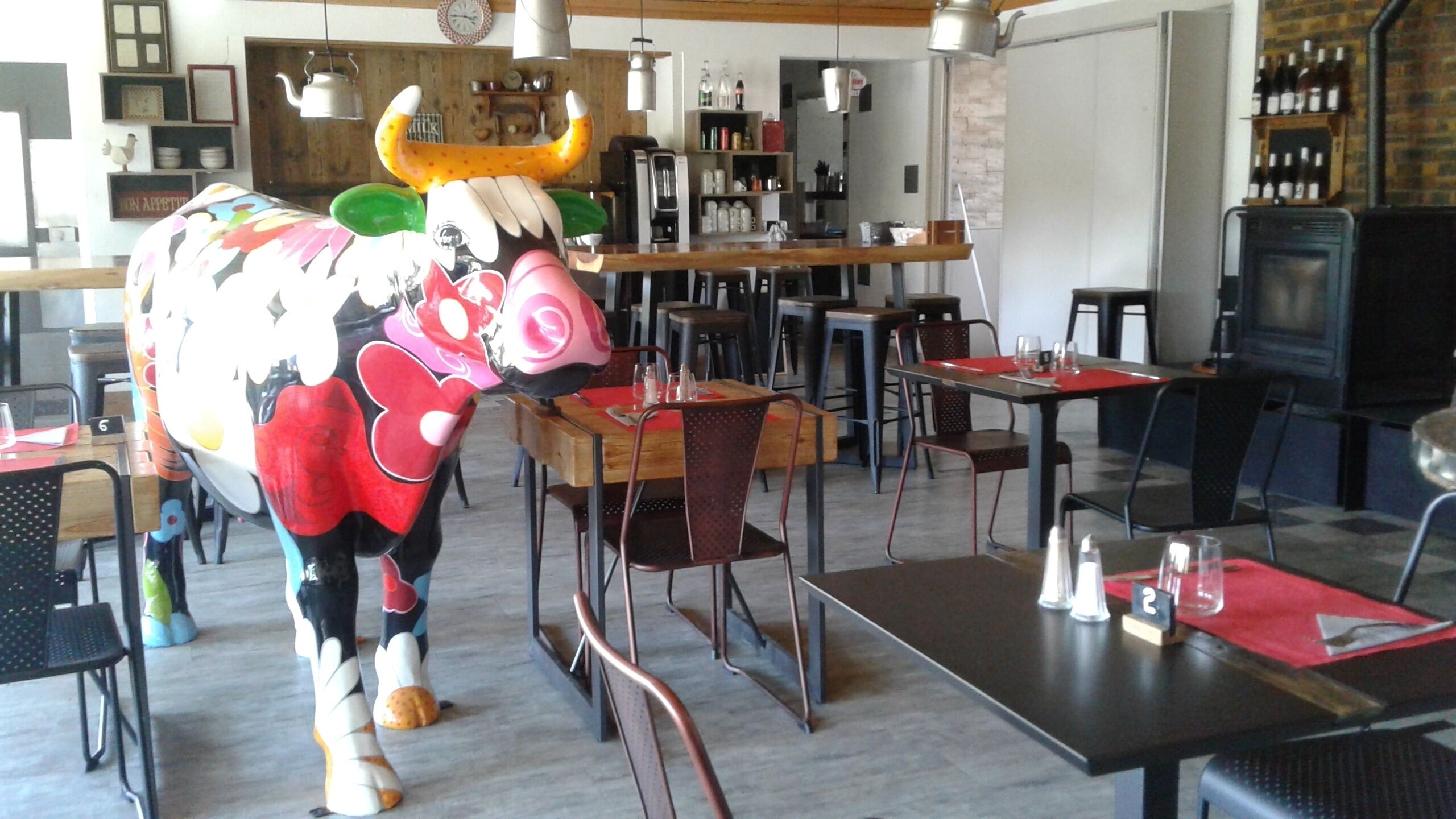 A restaurant interior with a large bull sculpture, wooden tables, metal chairs, and a brick wall.