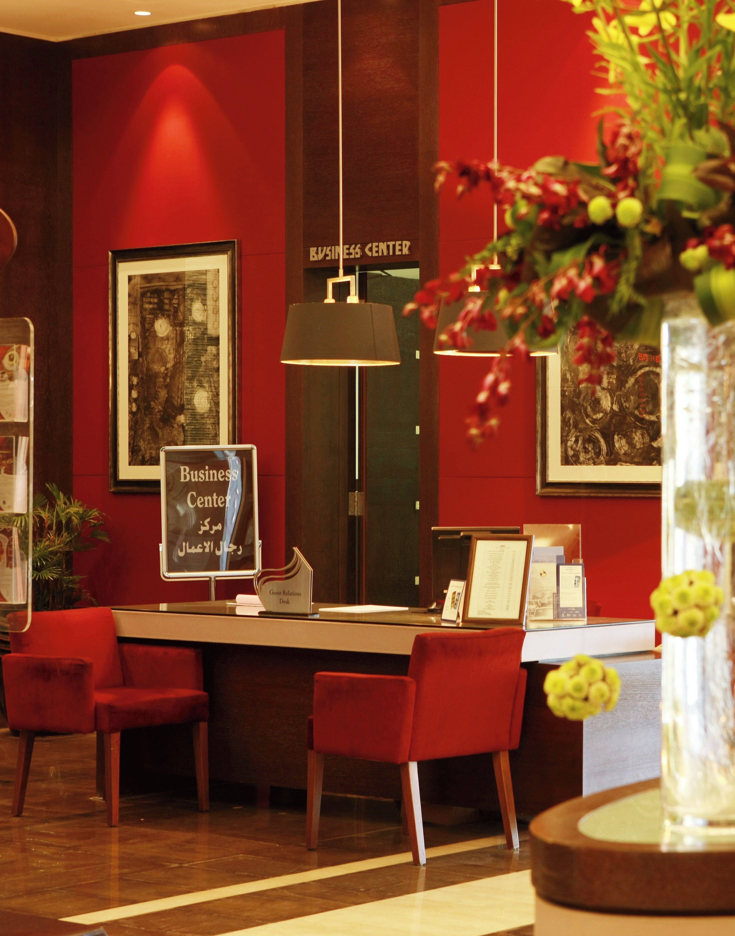 A reception desk with a sign indicating a guest relations desk, red chairs, a framed artwork, and a red wall with a business center sign.