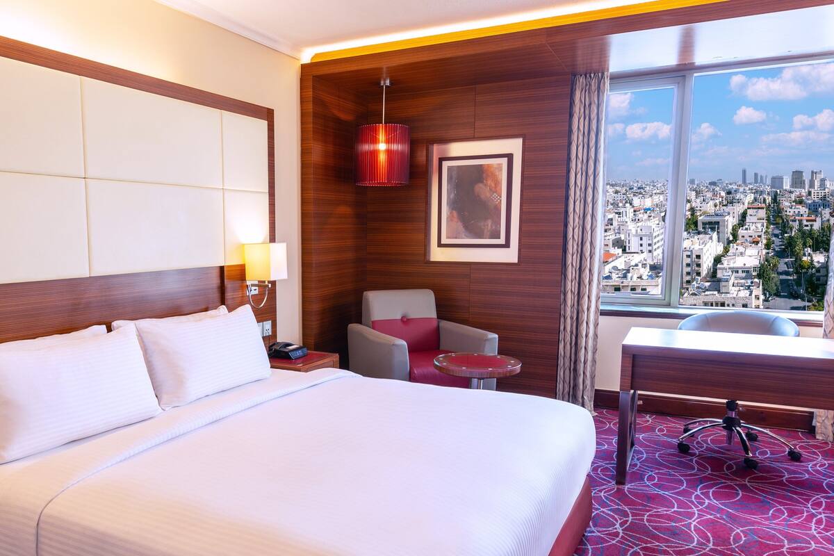 Habitación de hotel con una cama grande, un escritorio, una silla, una lámpara y vista a la ciudad por la ventana.