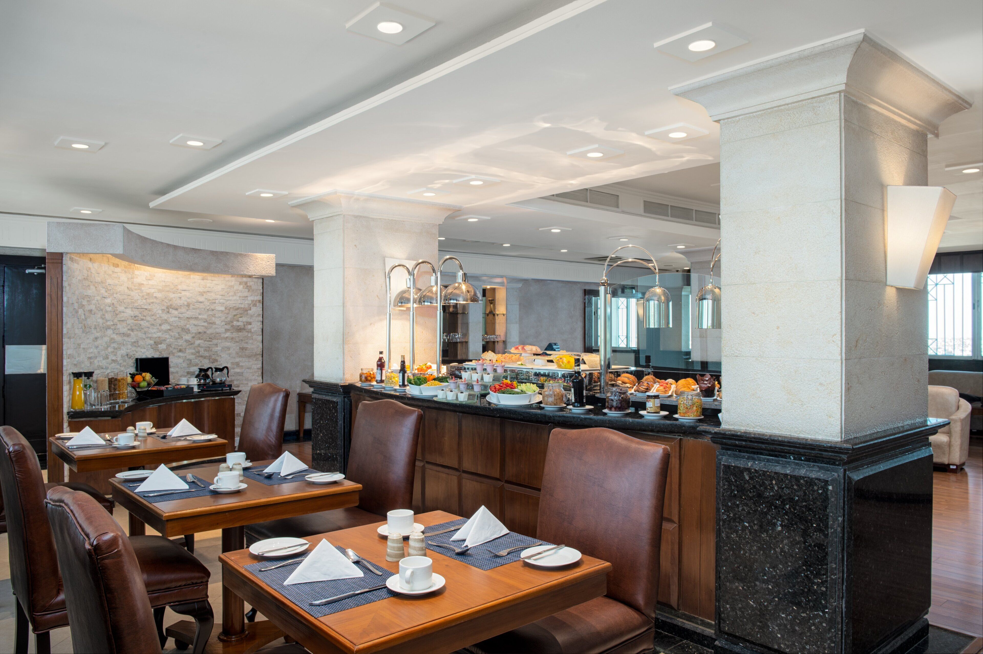 A modern dining area with wooden tables and leather chairs, a buffet counter with various food items, and a stone accent wall.