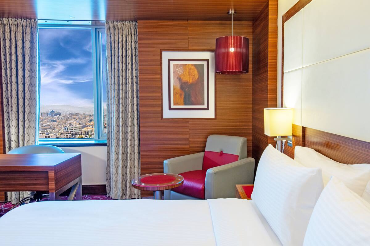 Habitación de hotel con cama, escritorio, silla, mesita, lámpara, un cuadro y vistas a la ciudad desde la ventana.