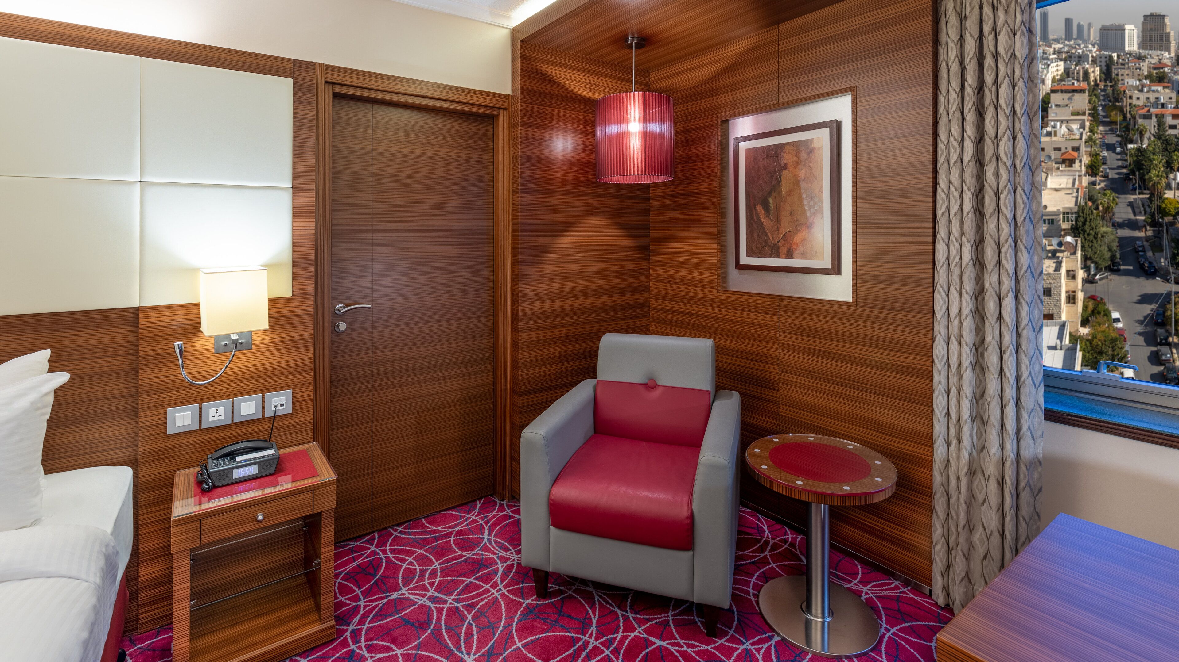 A hotel room with a wooden wardrobe, a red and grey armchair, a small bedside table with a lamp, a framed picture on the wall, and a city view through the window.