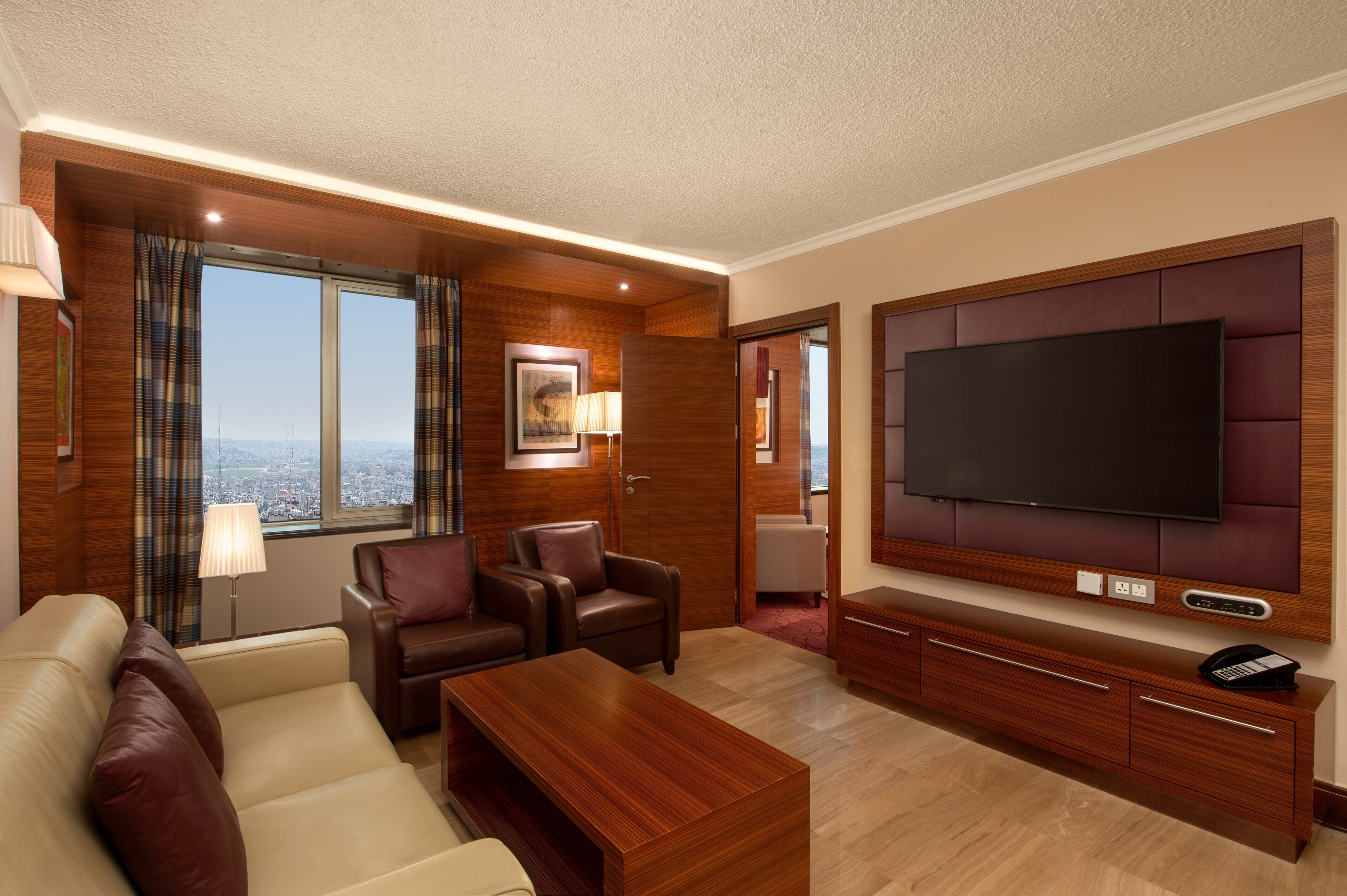 A hotel room with a large flat-screen TV, leather seating, a wooden coffee table, and a city view.