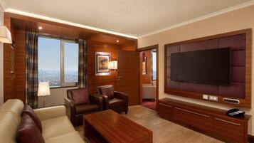 A hotel room with a large flat-screen TV, leather seating, a wooden coffee table, and a city view.