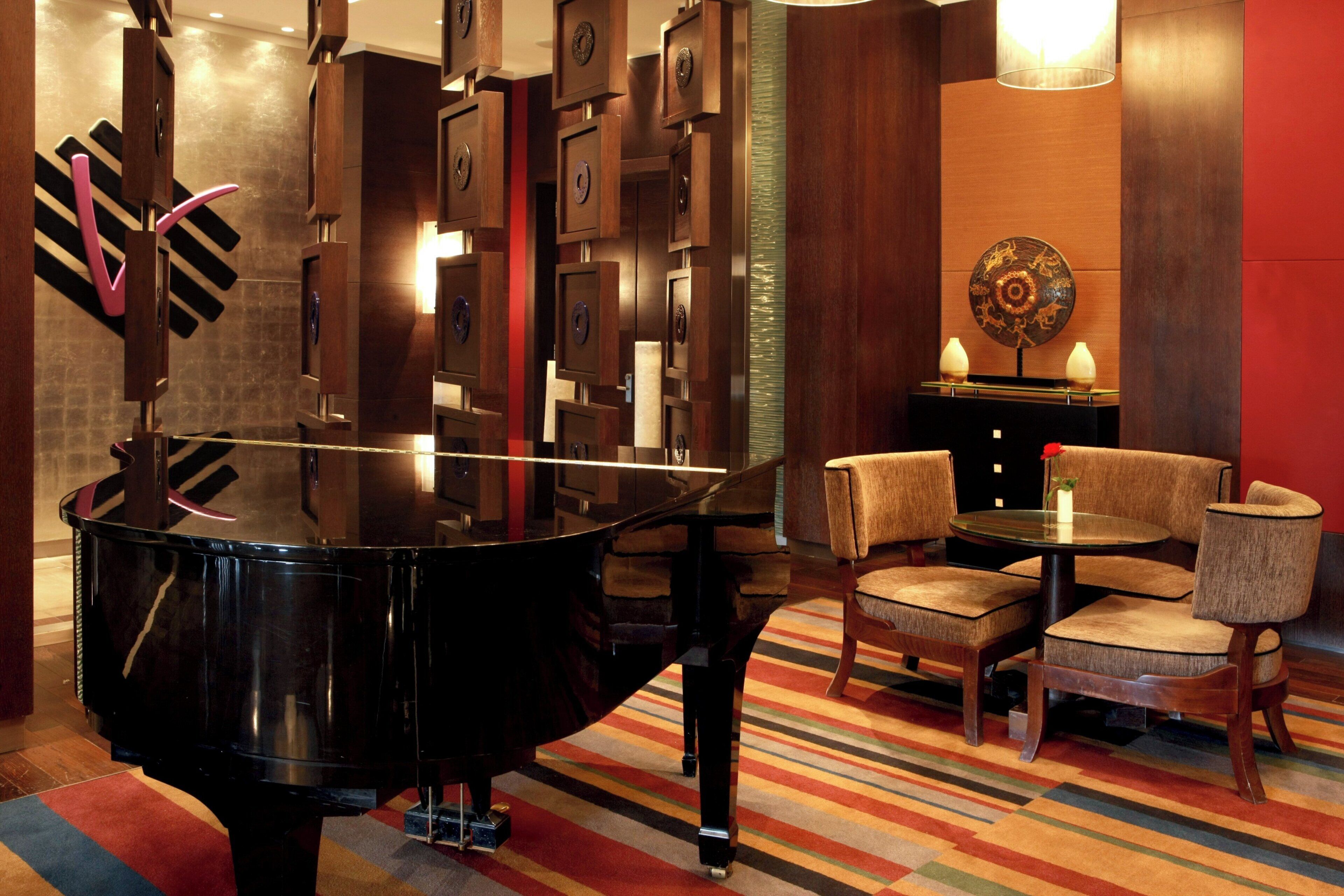 A grand piano in a modern room with a striped carpet, wooden chairs, and a small table with decorative items.