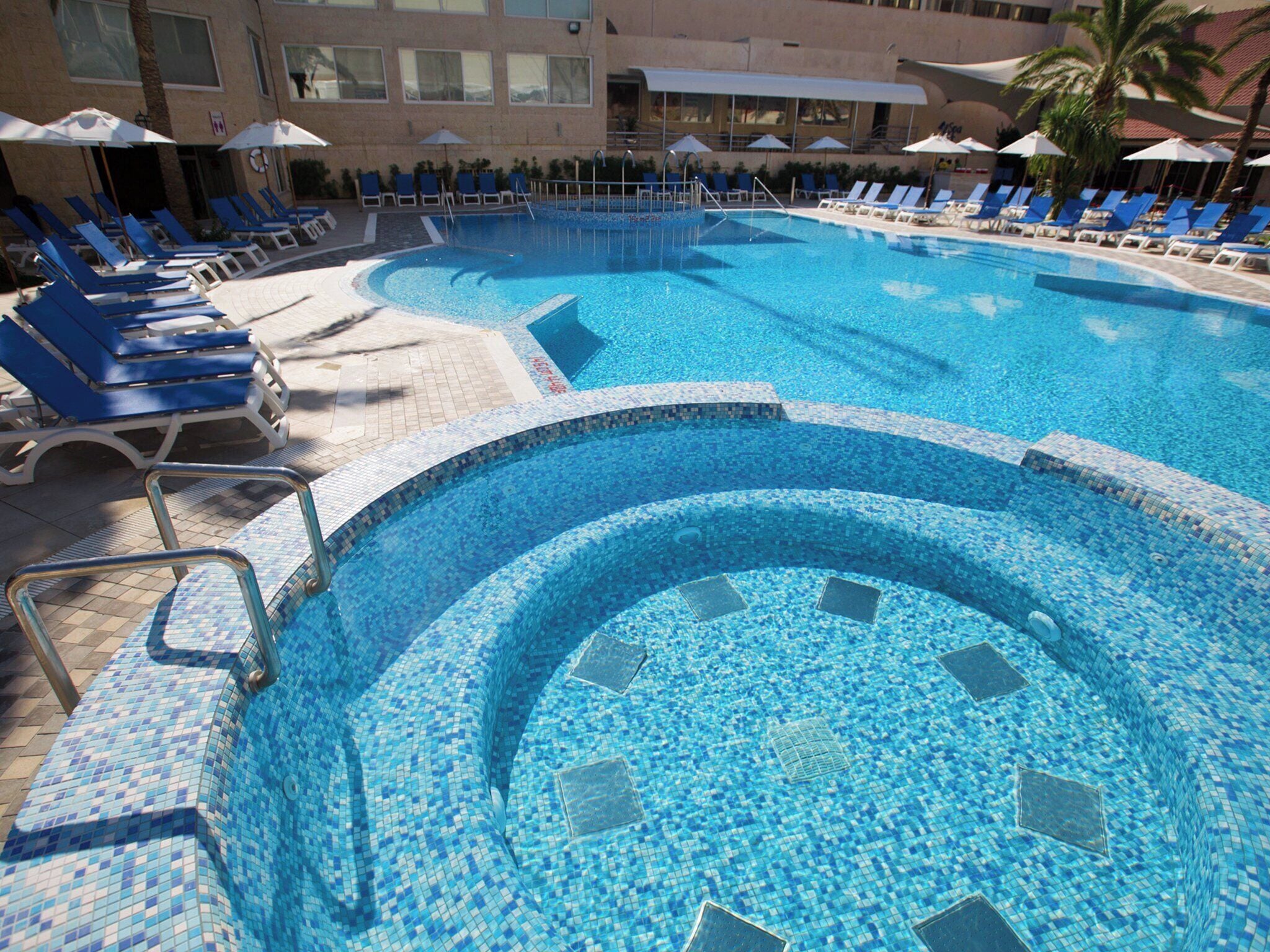 Una piscina con mosaicos azules, rodeada de sillones y sombrillas.