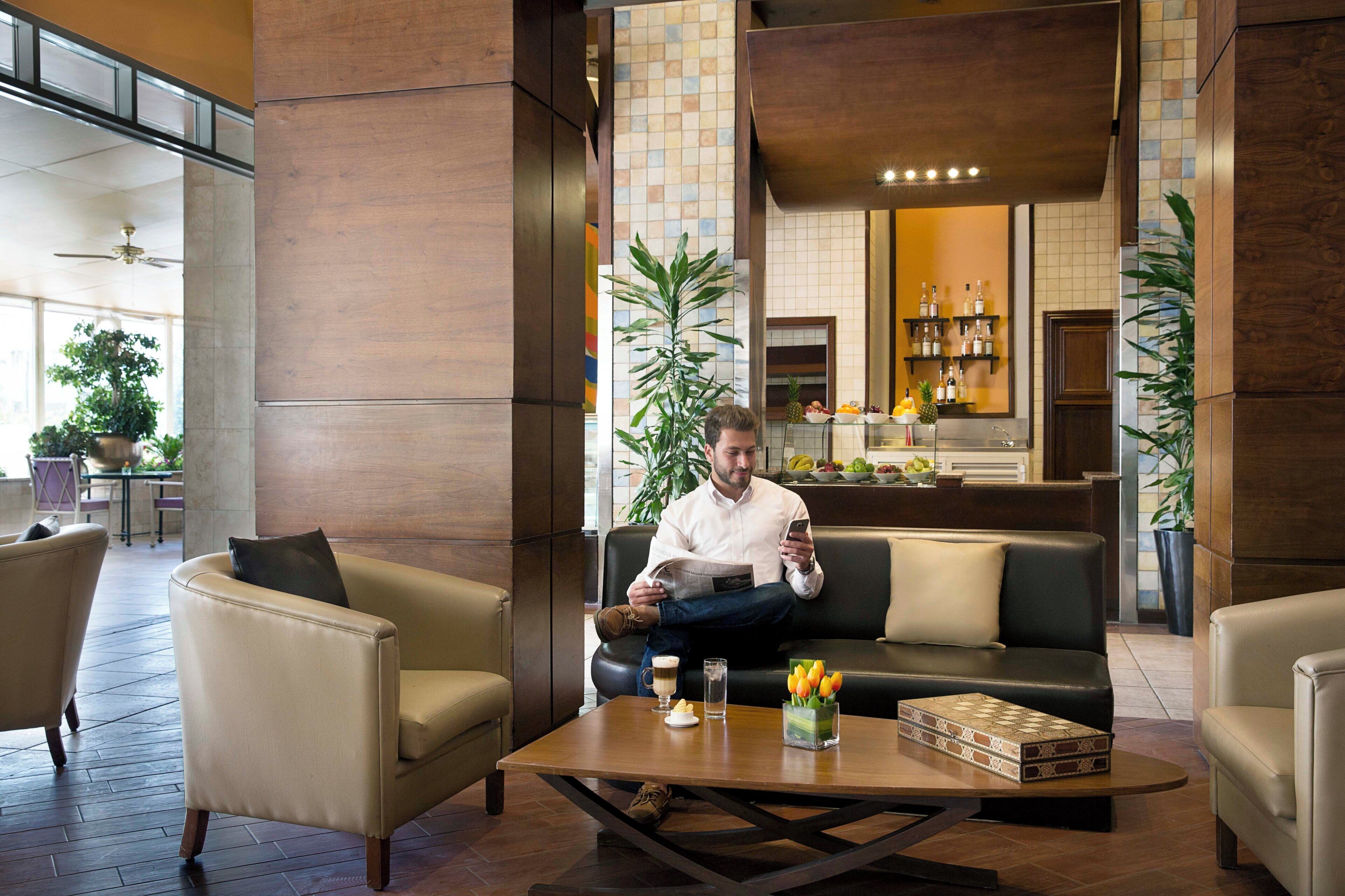 Een man zit op een bank in een moderne lobby, met een bijzettafeltje, een plant en een presentatie van fruit op de achtergrond.