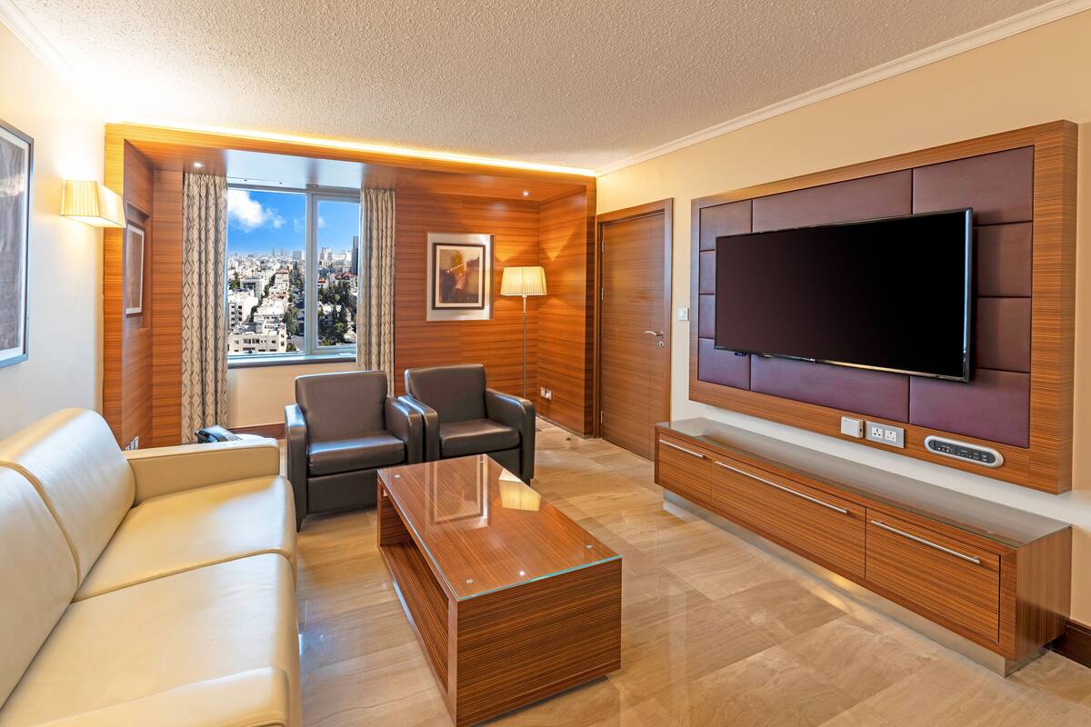 Una sala moderna con un sofá, dos sillones, una mesa de centro, un televisor y vistas a la ciudad a través de amplios ventanales.