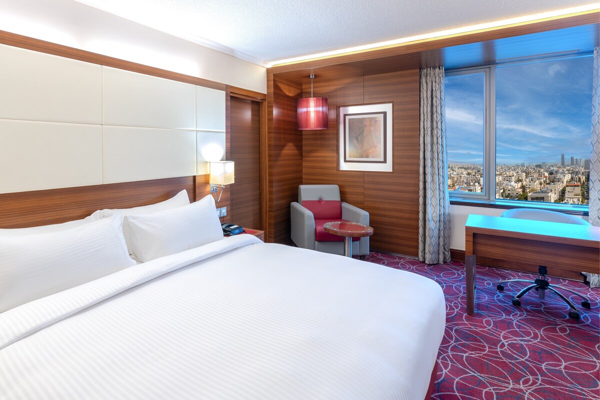 A hotel room with a large bed, a desk, a chair, and a view of a cityscape through a large window.