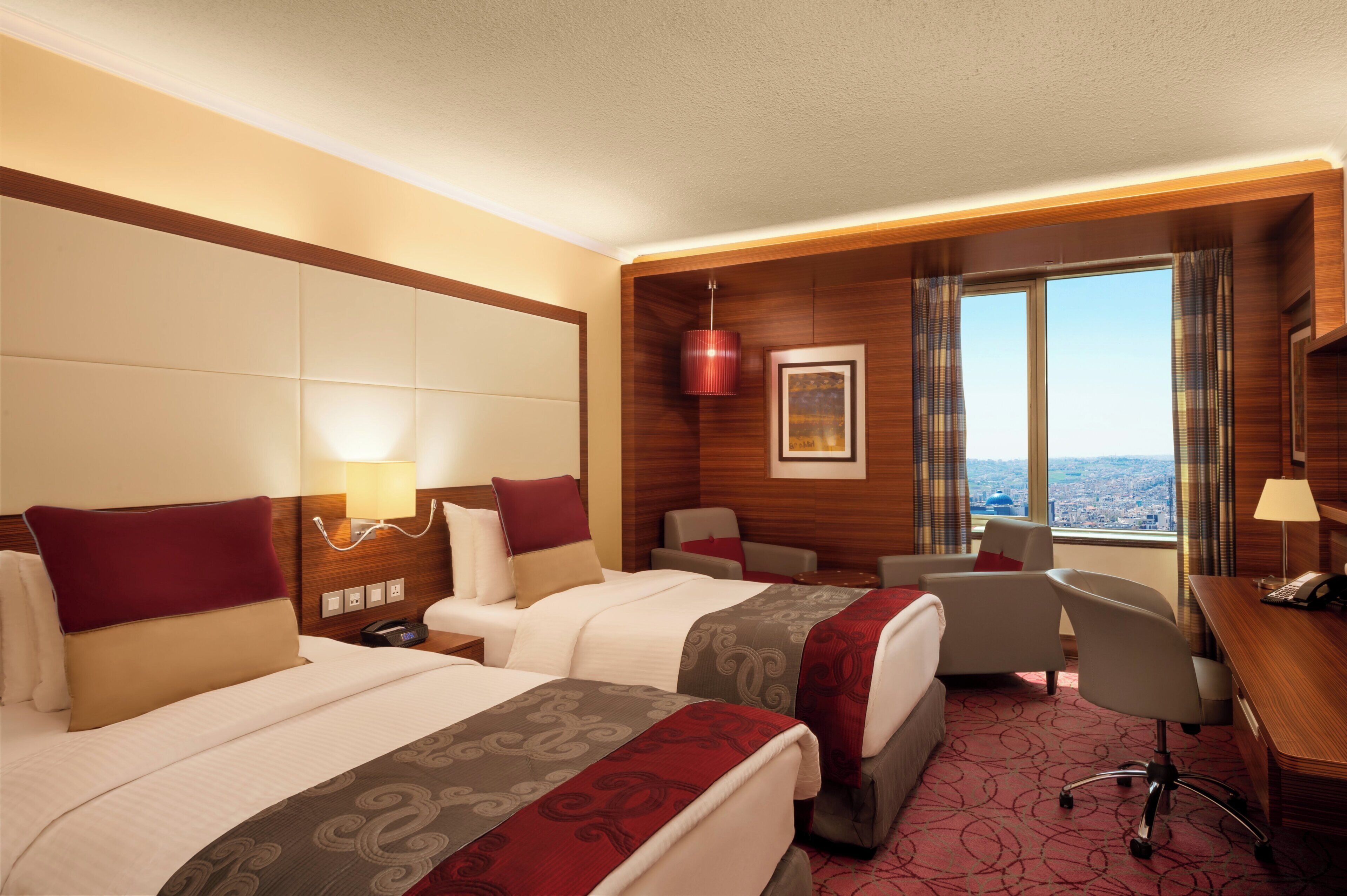 A hotel room with two beds, a desk, a chair, a window with a city view, and a framed picture on the wall.