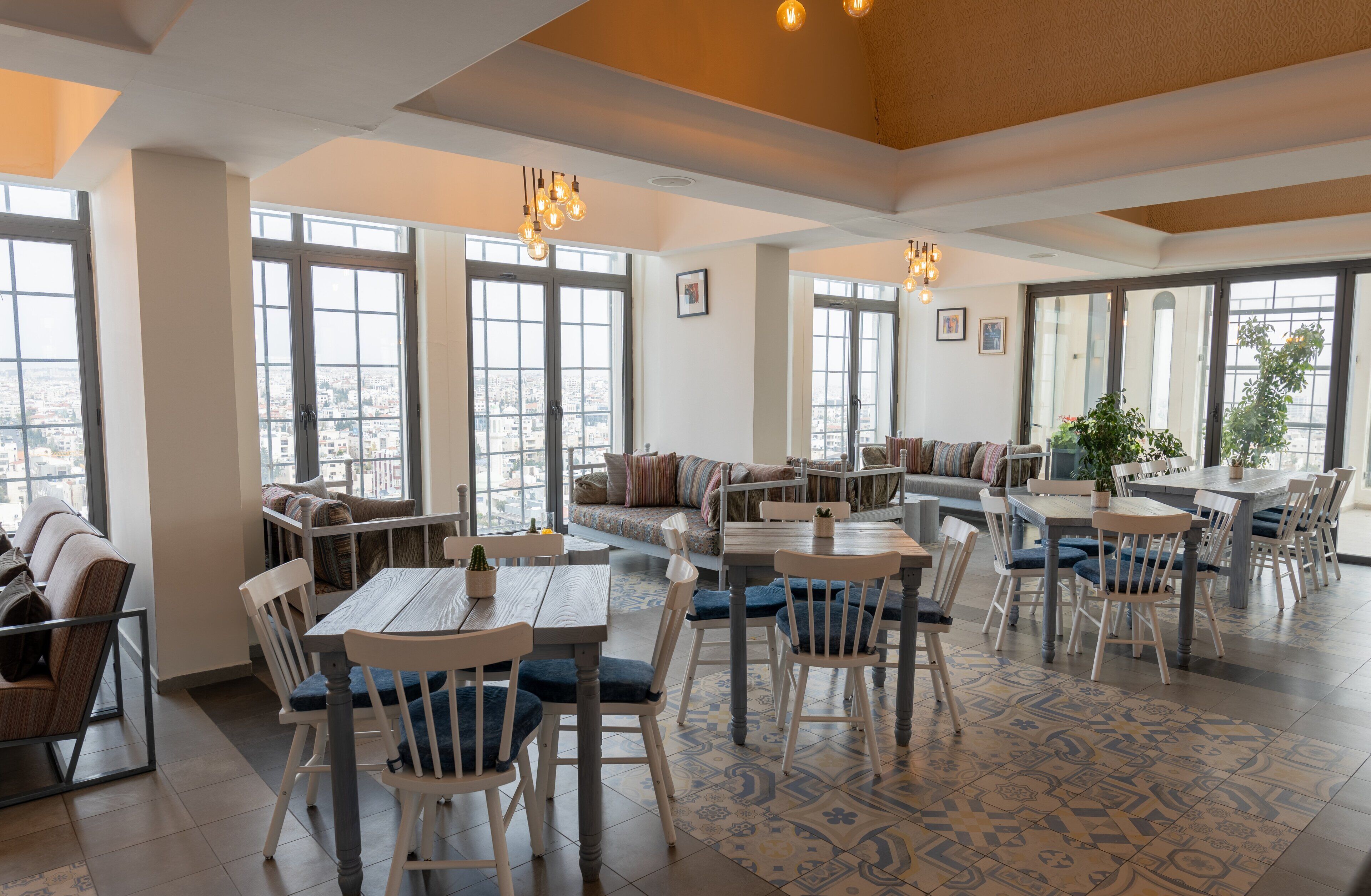 Uma sala de jantar ampla com pé direito alto, janelas grandes e diversos tipos de assentos, incluindo mesas e sofás.