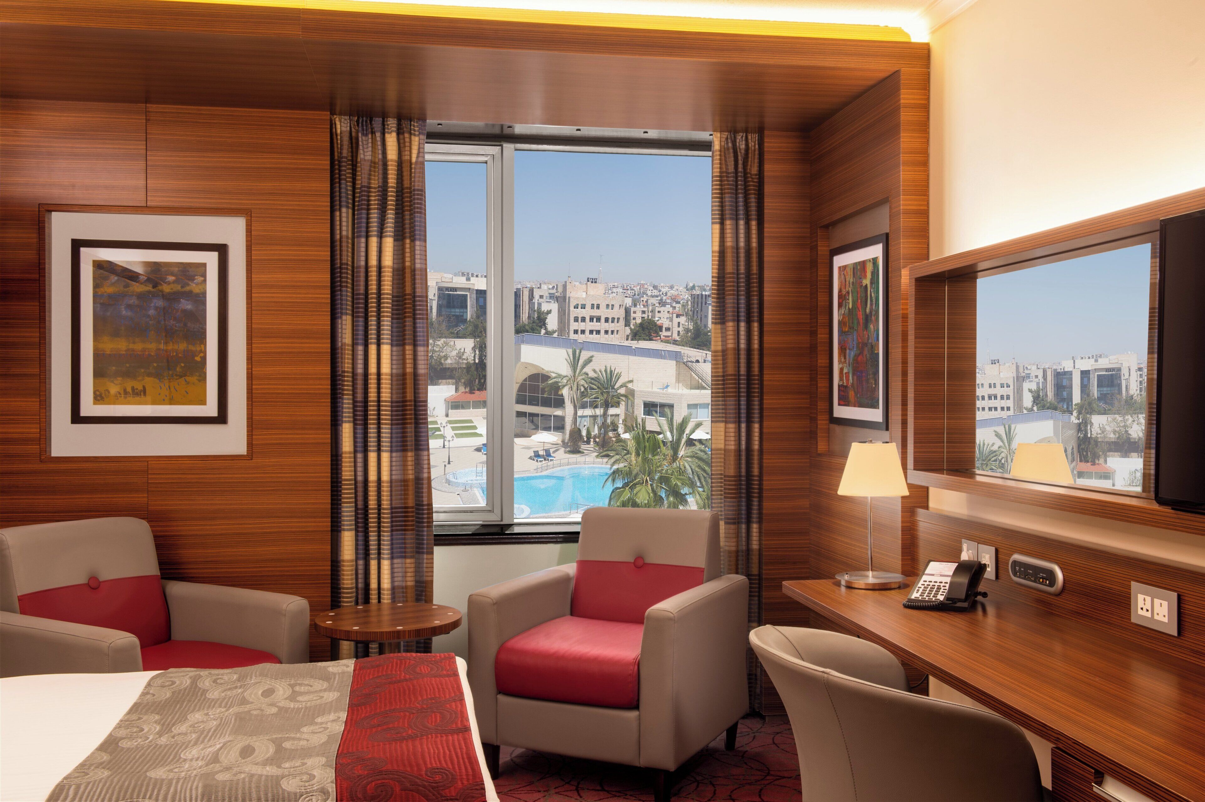 Ein Hotelzimmer mit einem Bett, zwei Sesseln, einem Schreibtisch mit Telefon und einem großen Fenster mit Blick auf die Stadt.