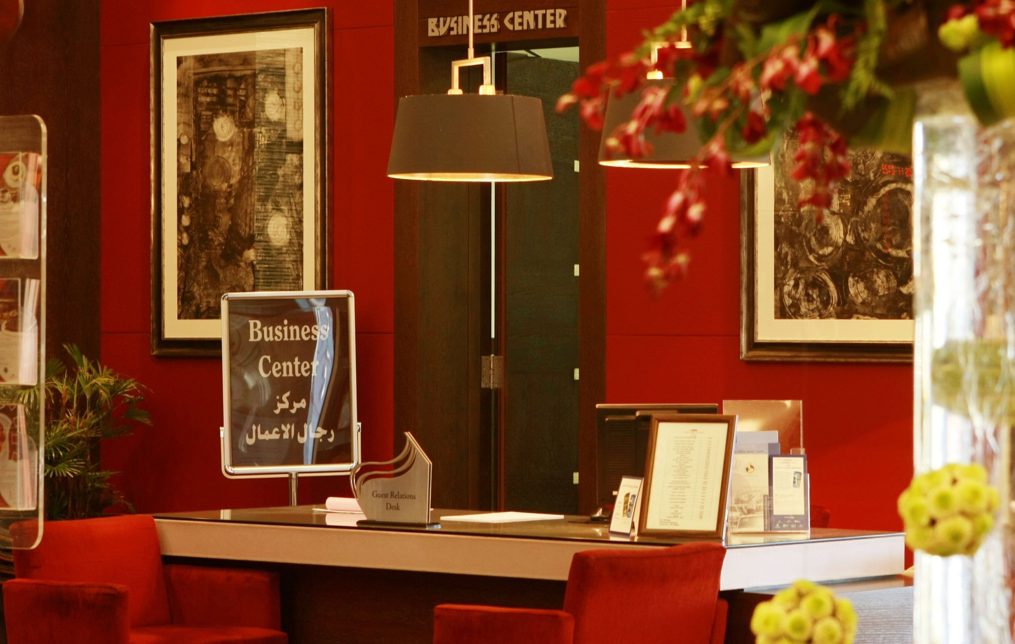A reception desk with a sign reading "Business Center" and "Guest Relations Desk," red chairs, a black pendant light, framed artwork, and a potted plant.
