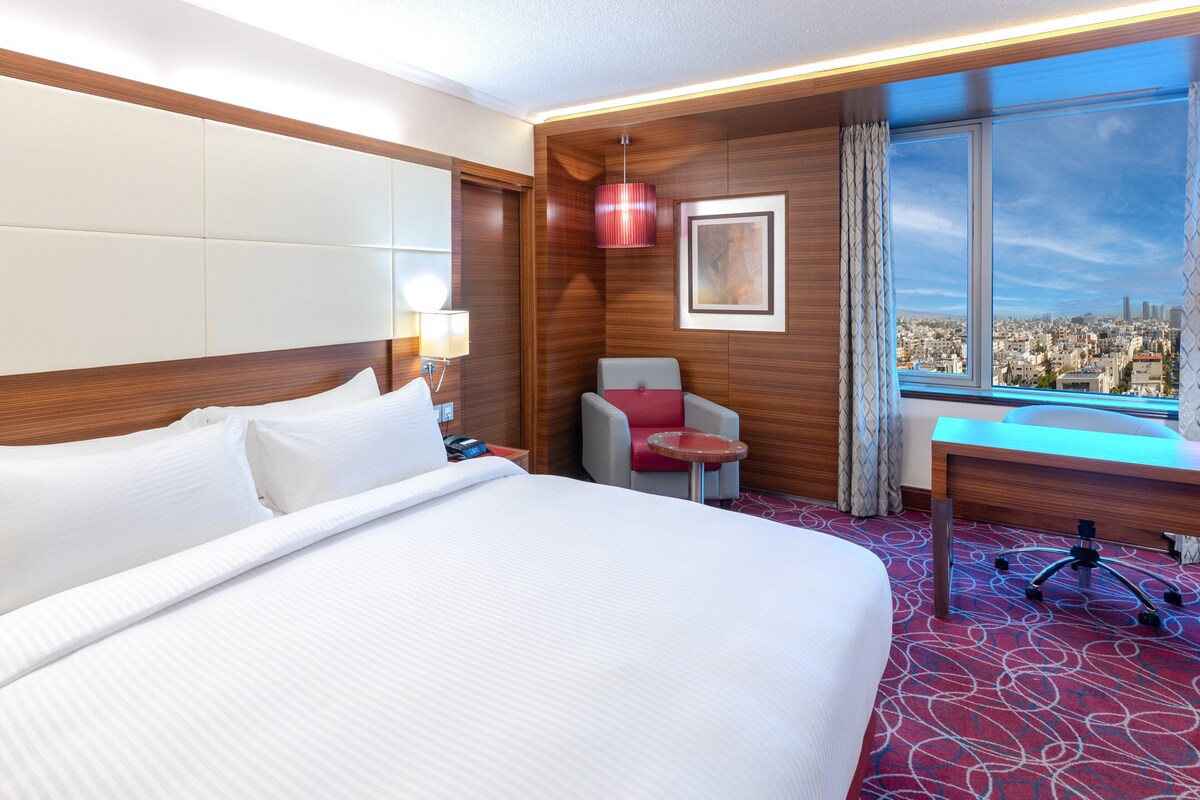 A hotel room with a large bed, a desk, a chair, and a view of a cityscape through a large window.