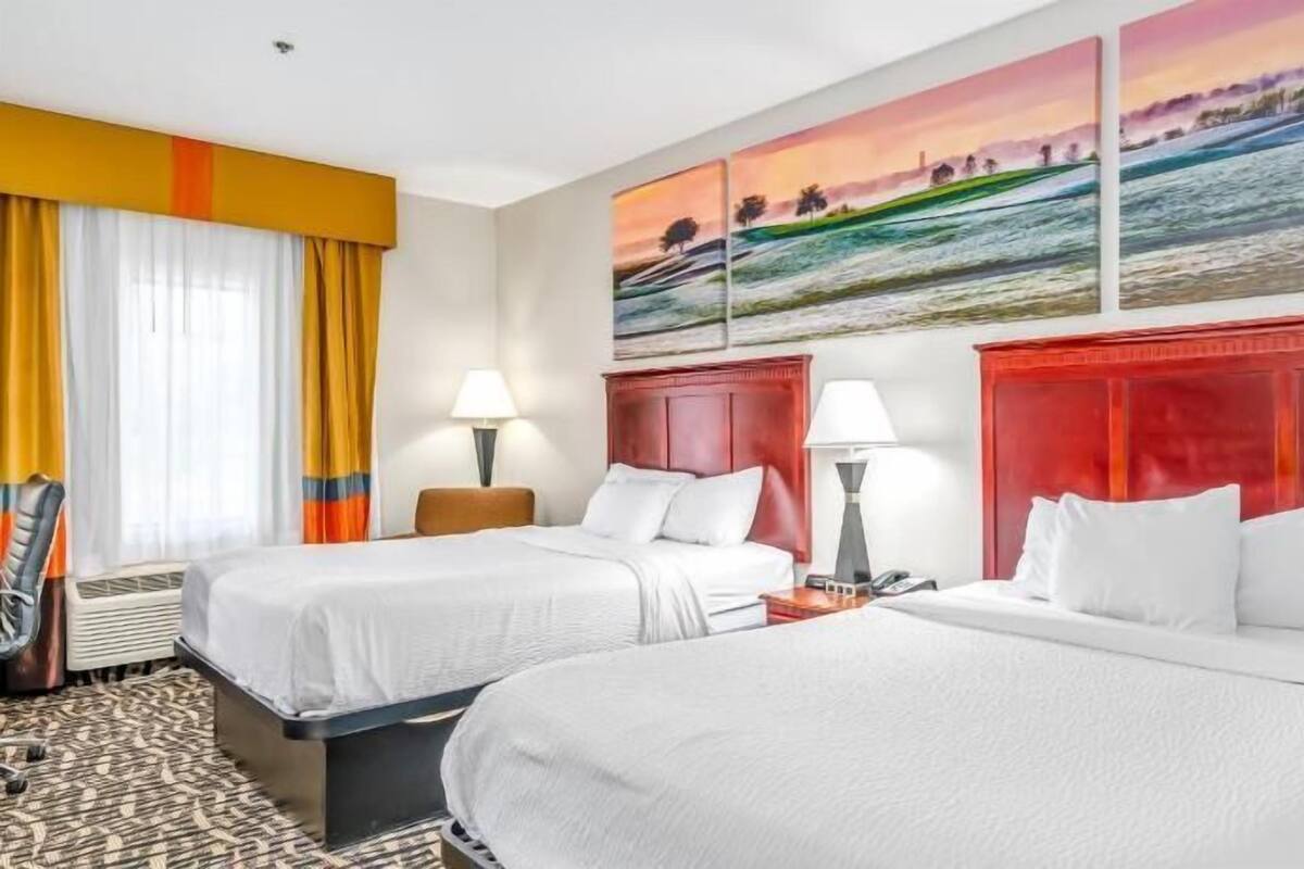 Habitación de hotel con dos camas, cabecera roja, un cuadro de un paisaje y una ventana con cortinas.