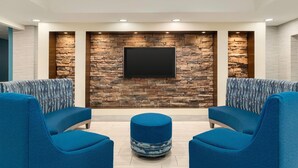 A modern lounge area with a stone wall, a flat-screen TV, and blue upholstered seating. - Days Inn & Suites by Wyndham Prattville-Montgomery (Prattville)