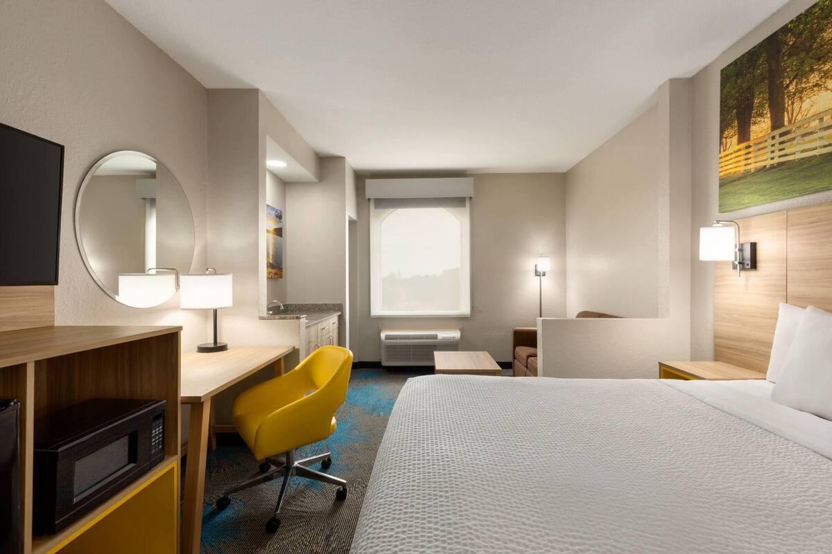 Habitación de hotel con cama, escritorio, silla, microondas, lámpara y un ventanal grande.