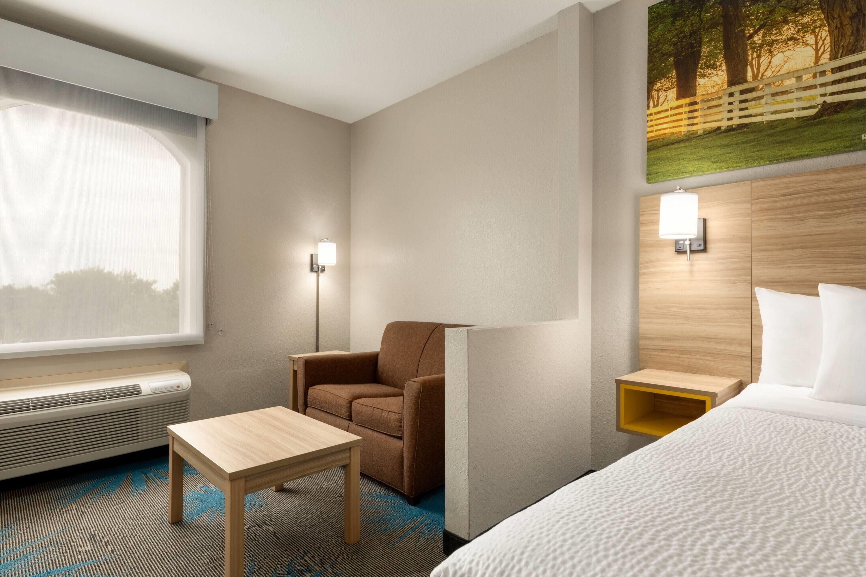 Una habitación de hotel con una cama, una silla, una mesita, una lámpara de pared y un cuadro de un paisaje boscoso.