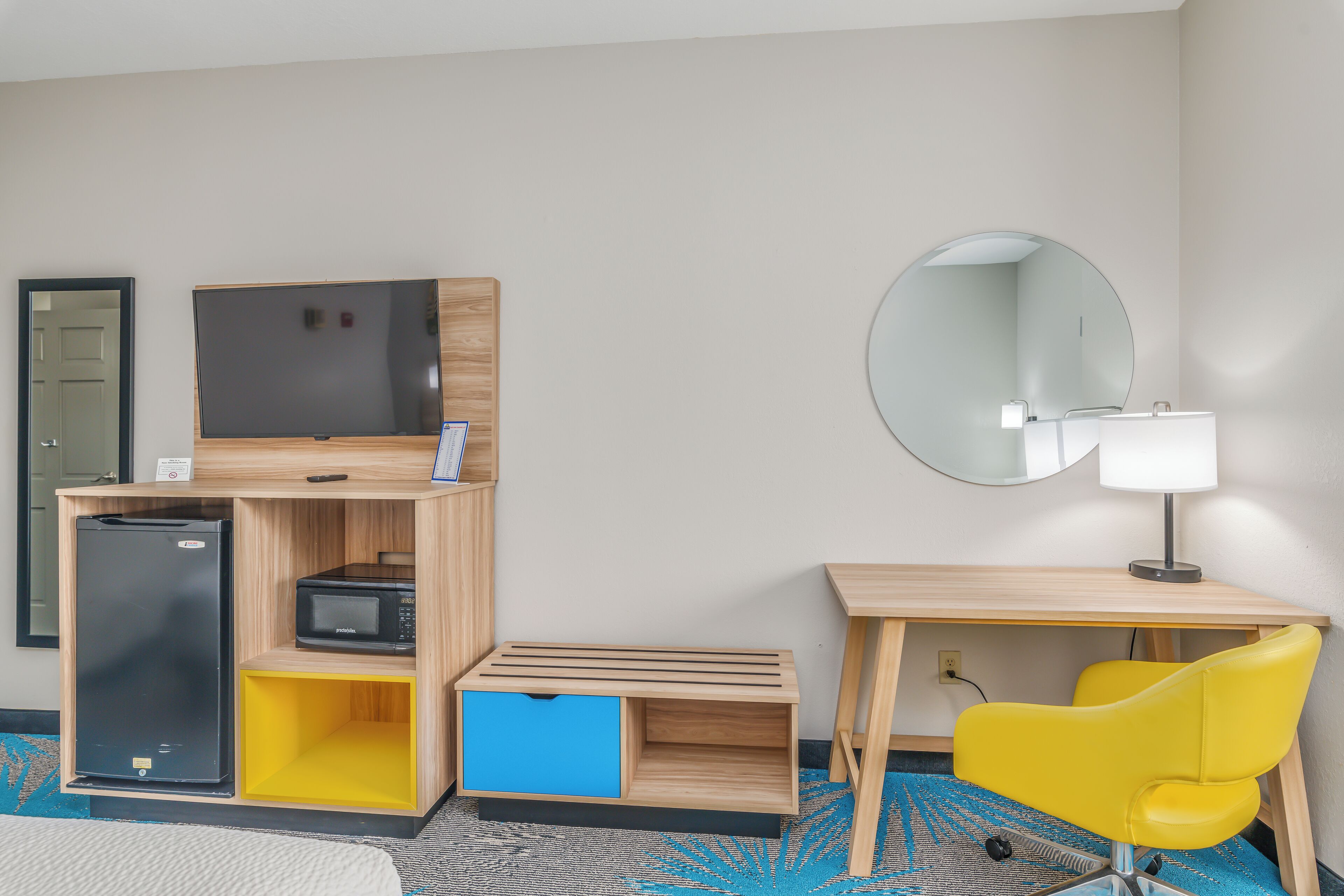 Quarto de hotel compacto com cama, escrivaninha de madeira com cadeira amarela, televisão de tela plana, micro-ondas e pequeno refrigerador.