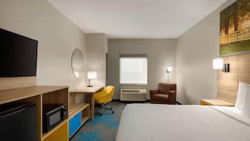 Una habitación de hotel con cama, escritorio y silla, televisor, aire acondicionado y un cuadro en la pared.