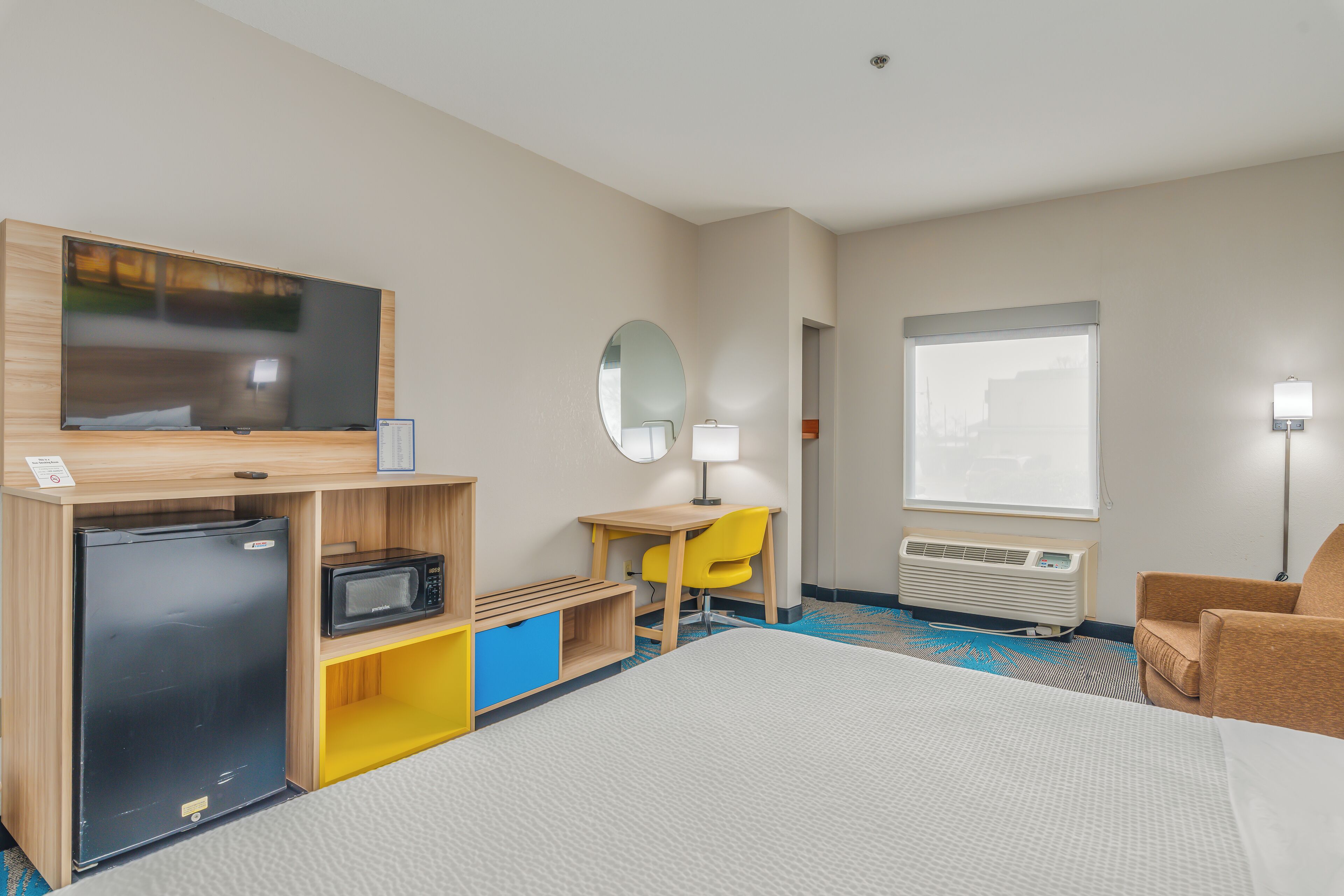 A hotel room with a bed, a desk with a chair, a television, a microwave, a refrigerator, a mirror, a window, and a lamp.