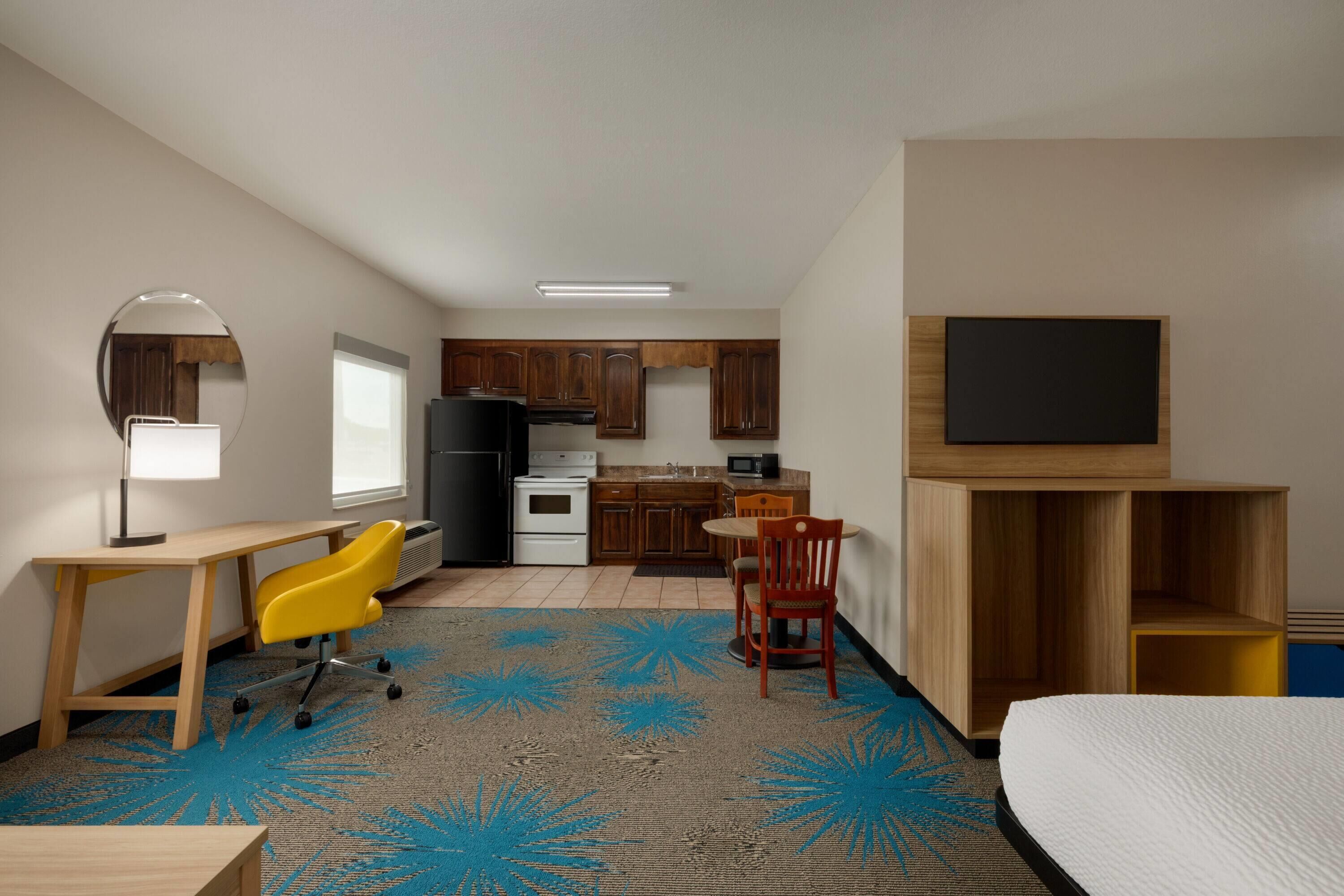 Een kleine hotelkamer met kitchenette, bureau met stoel, bed, televisie en spiegel.