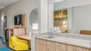 A modern hotel room with a wooden desk, a yellow chair, a large mirror, a sink, and a television. - Days Inn & Suites by Wyndham Prattville-Montgomery (Prattville)