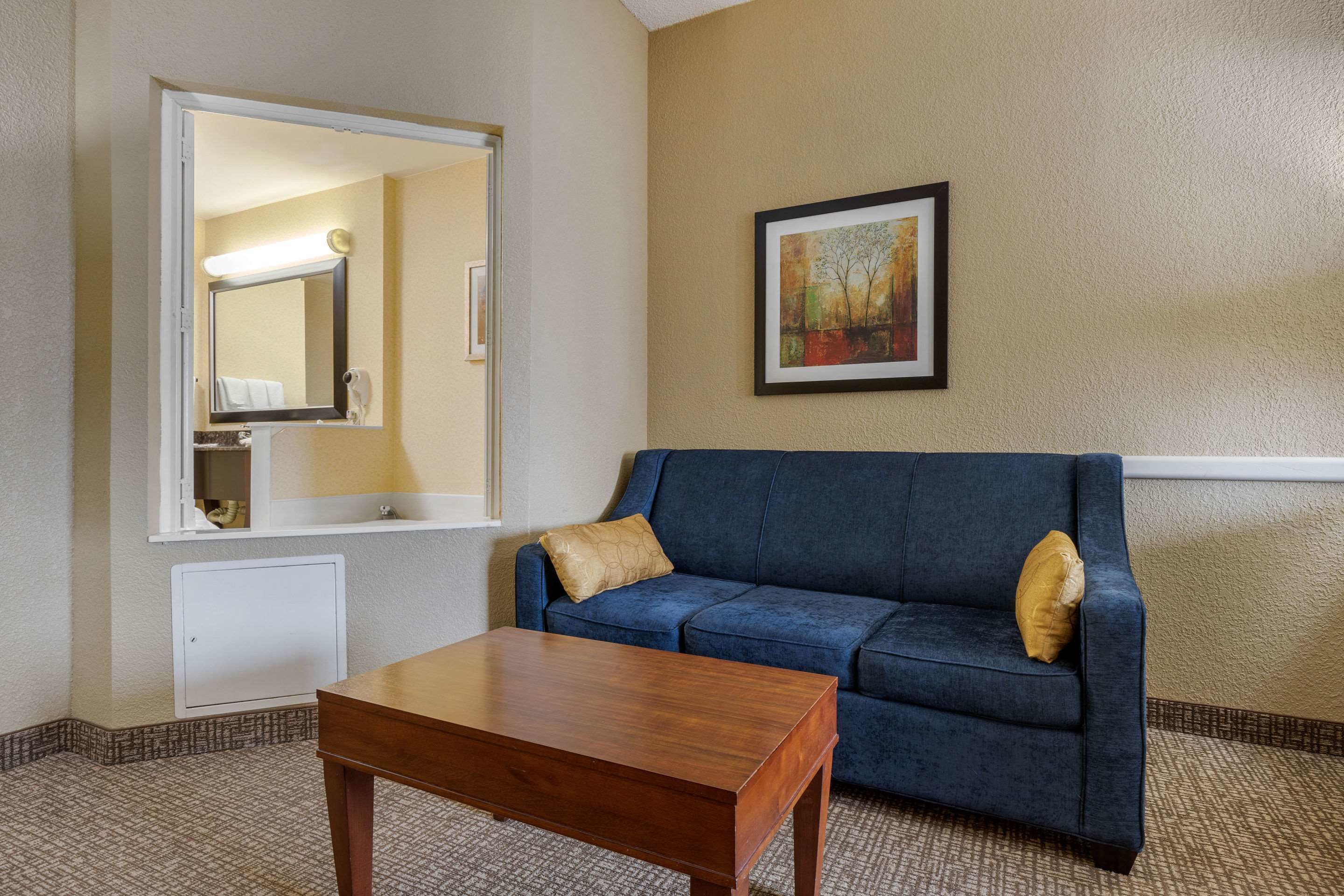 A hotel room with a blue sofa, a wooden coffee table, a large mirror, a framed painting, and a white cabinet.