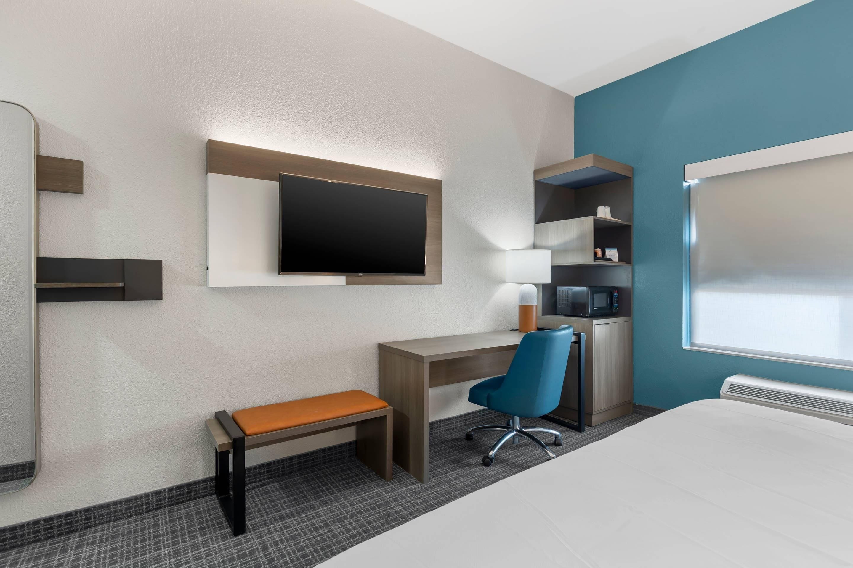 Habitación de hotel con cama, escritorio con silla, televisor, microondas y una pequeña kitchenette.