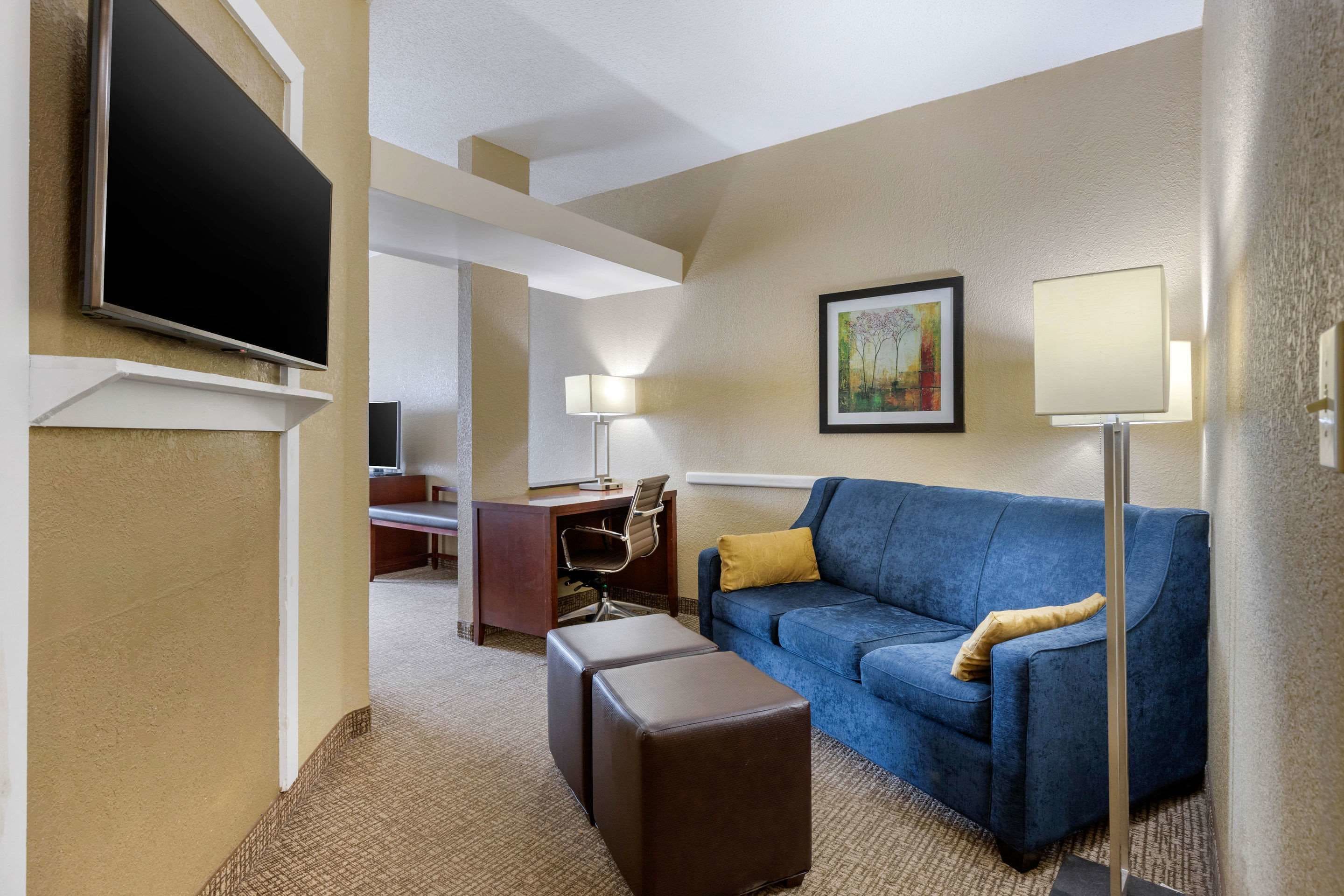A hotel room with a blue sofa, a flat-screen TV mounted on the wall, a desk with a chair, and a lamp.