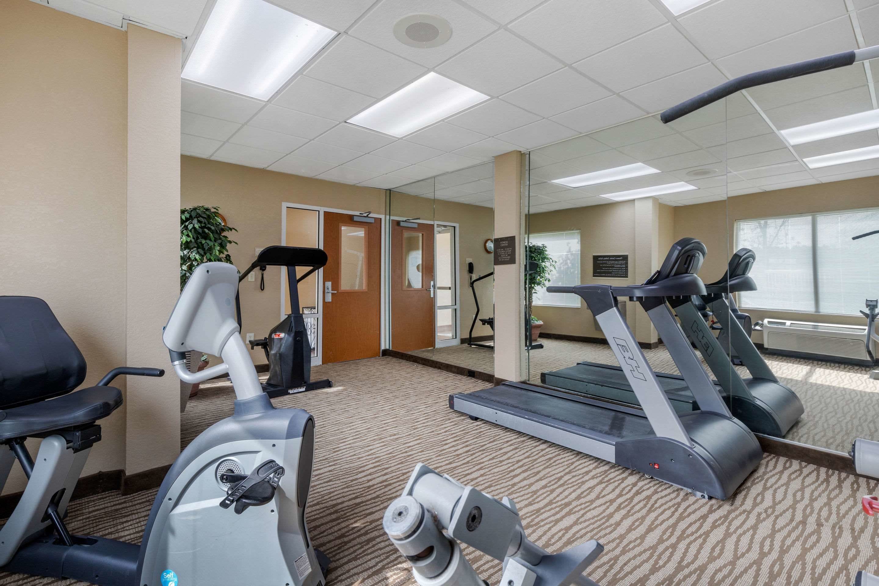 A fitness center with exercise equipment, including treadmills and an elliptical machine, carpeted flooring, and a large mirror reflecting the space.