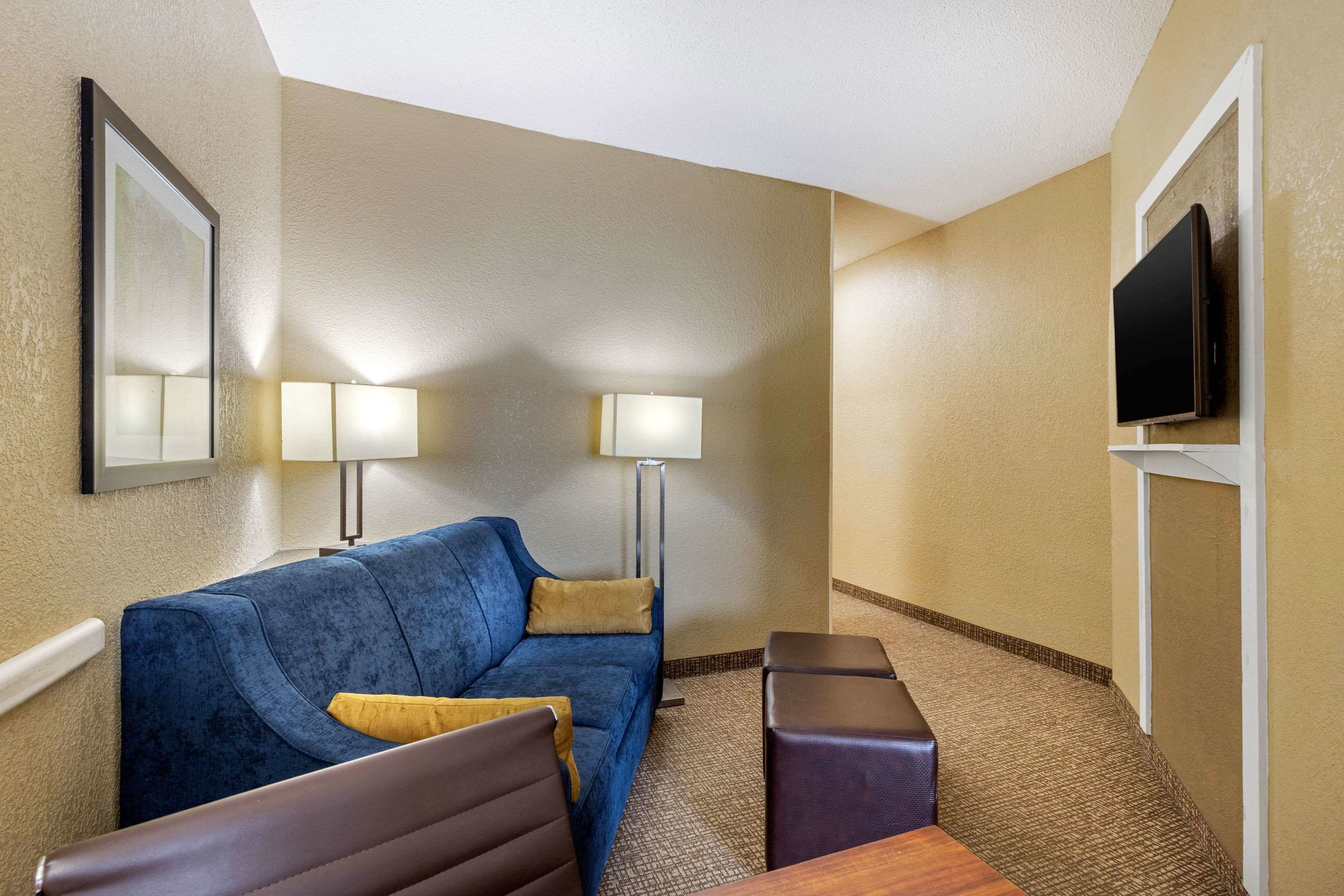 Habitación de hotel con sofá azul, dos mesitas auxiliares, un televisor de pantalla plana montado en la pared y dos lámparas de pie.