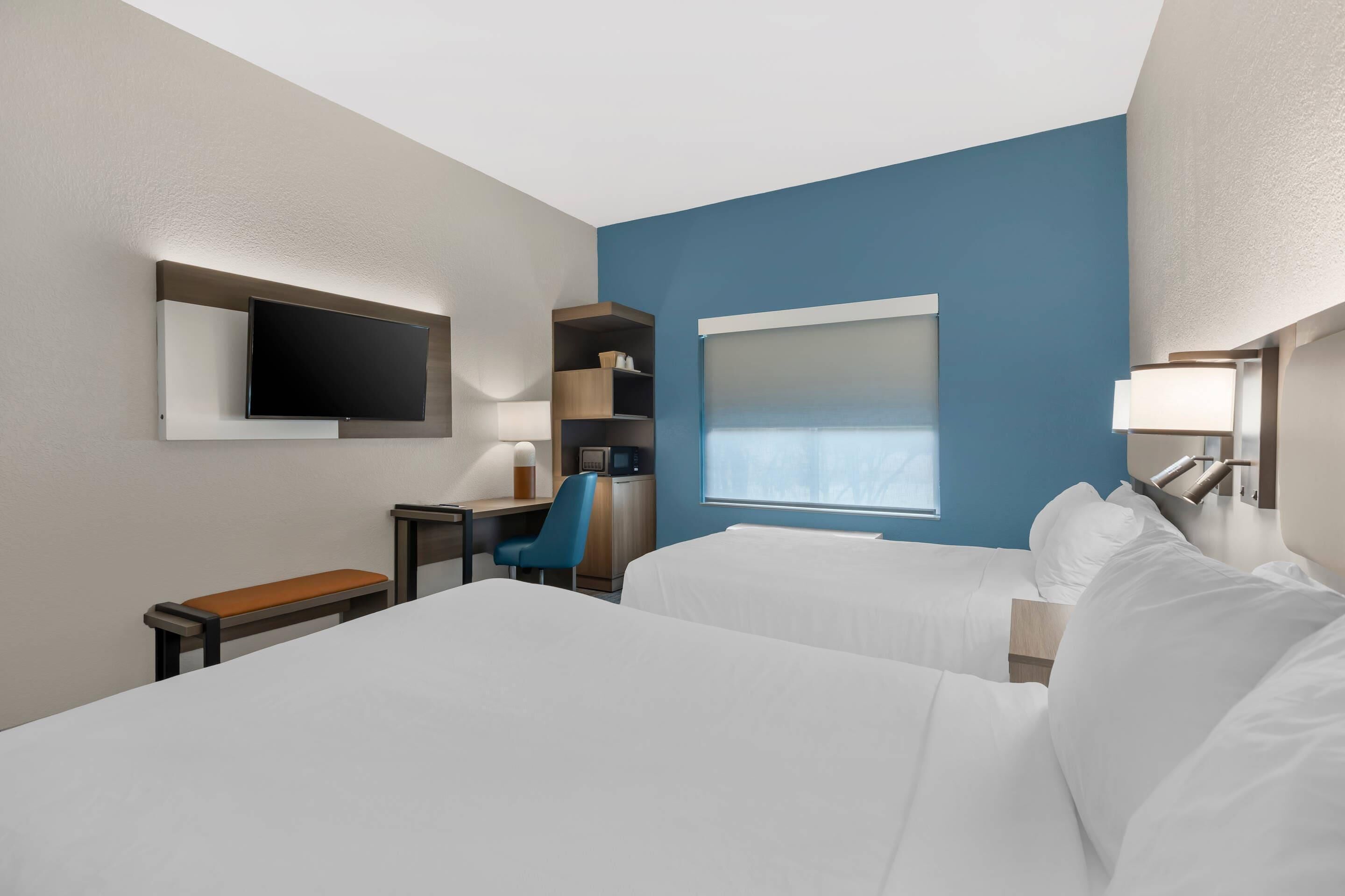 Een hotelkamer met twee bedden, een bureau met stoel, een televisie, een raam en een plank met spullen.