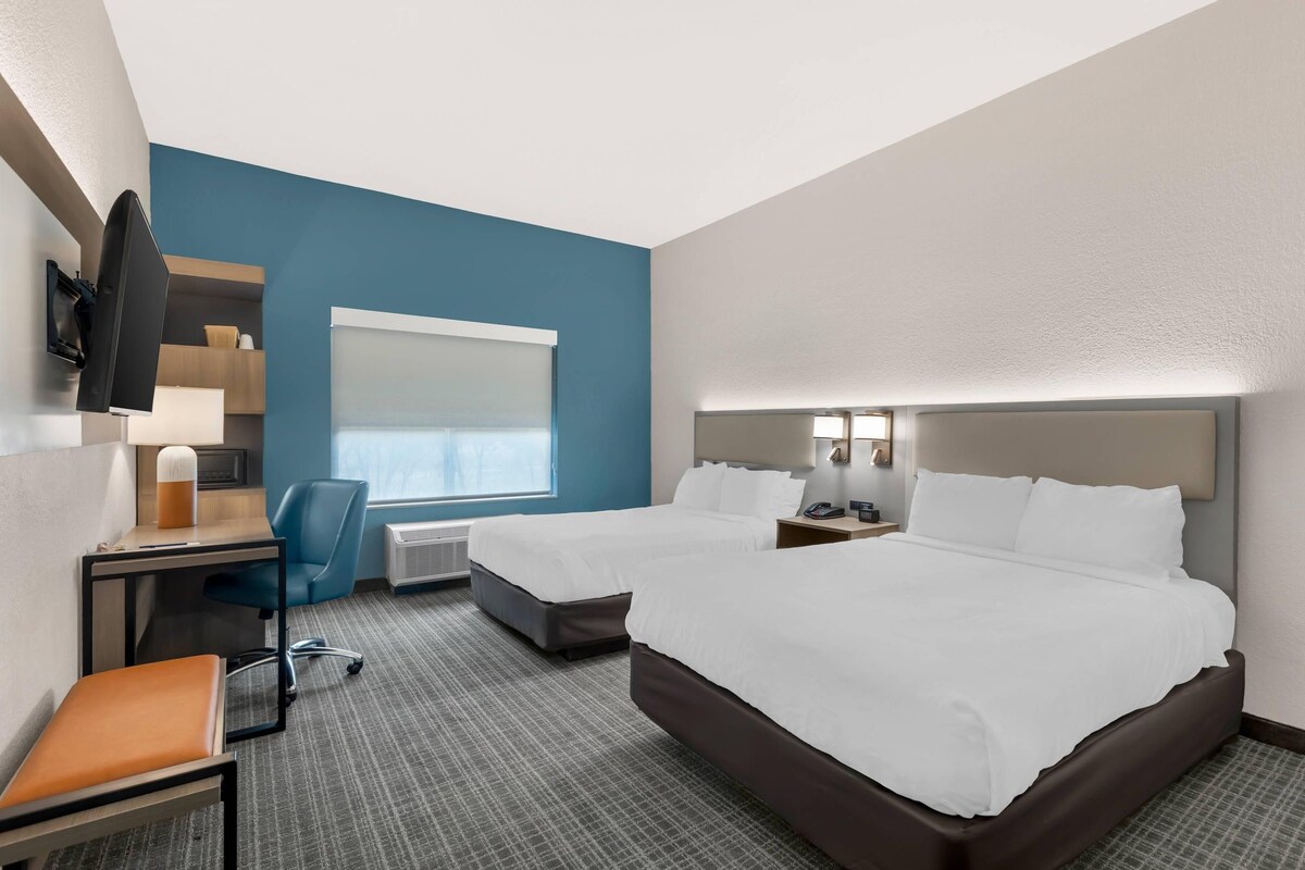 A hotel room with two beds, a desk with a chair, a TV mounted on the wall, and a window with blinds.