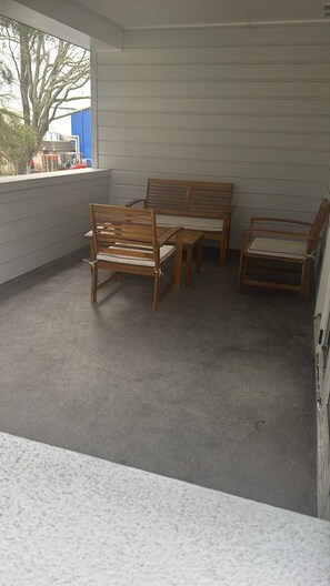 A covered outdoor seating area with a bench and chairs, a window with a view of trees and buildings, and a grey carpeted floor.