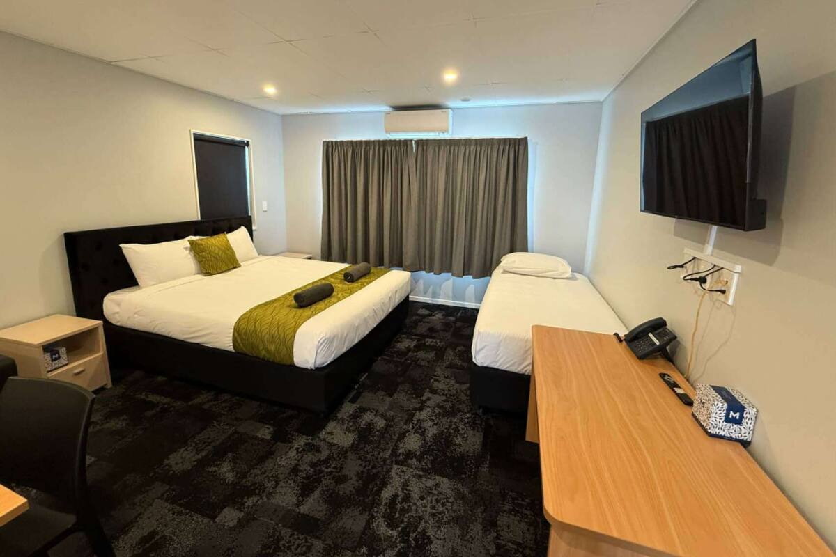 Una habitación de hotel con una cama grande, una cama más pequeña, un escritorio, una silla, un televisor y una ventana con cortinas.