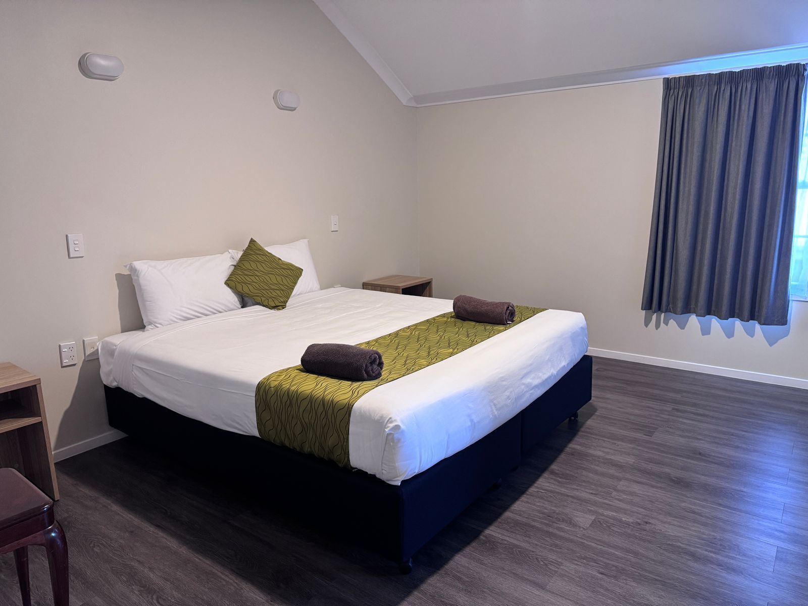 Una habitación de hotel con una cama, una mesita de noche, piso de madera y una ventana con cortinas.