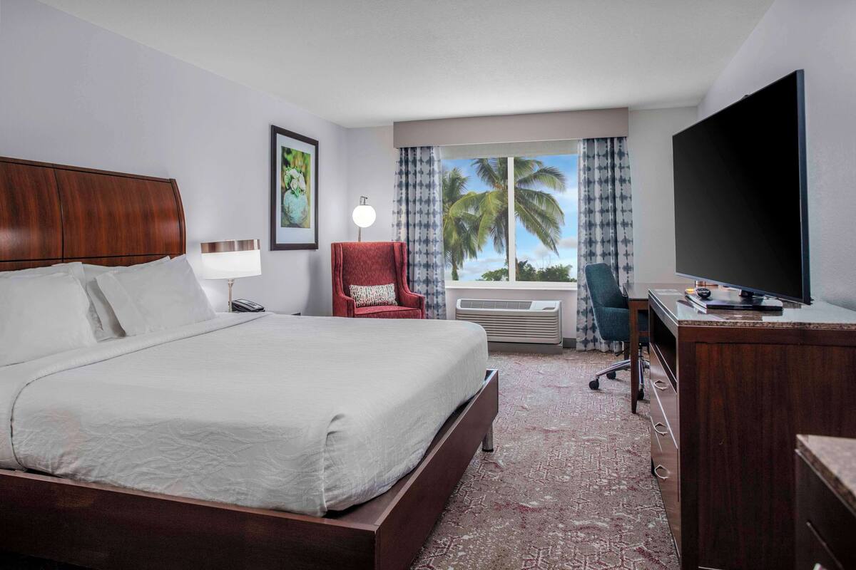 Une chambre d’hôtel avec un grand lit, un bureau, une chaise, une télévision et une fenêtre donnant sur des palmiers.
