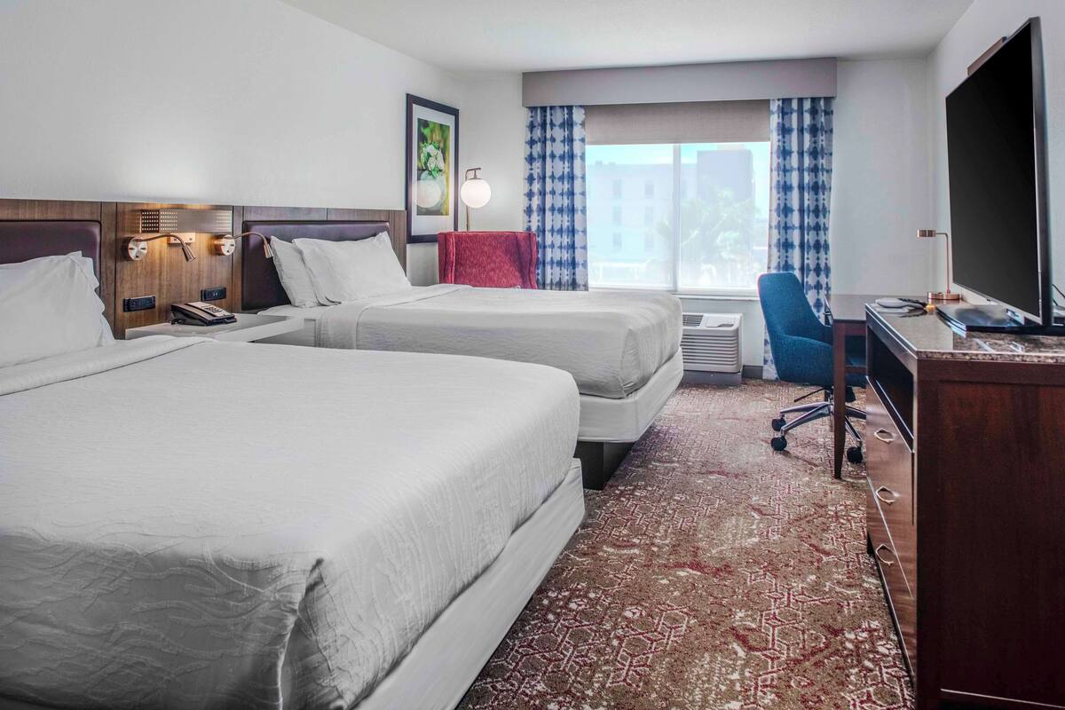 A hotel room with two beds, a desk, a chair, a TV, and a window with curtains.
