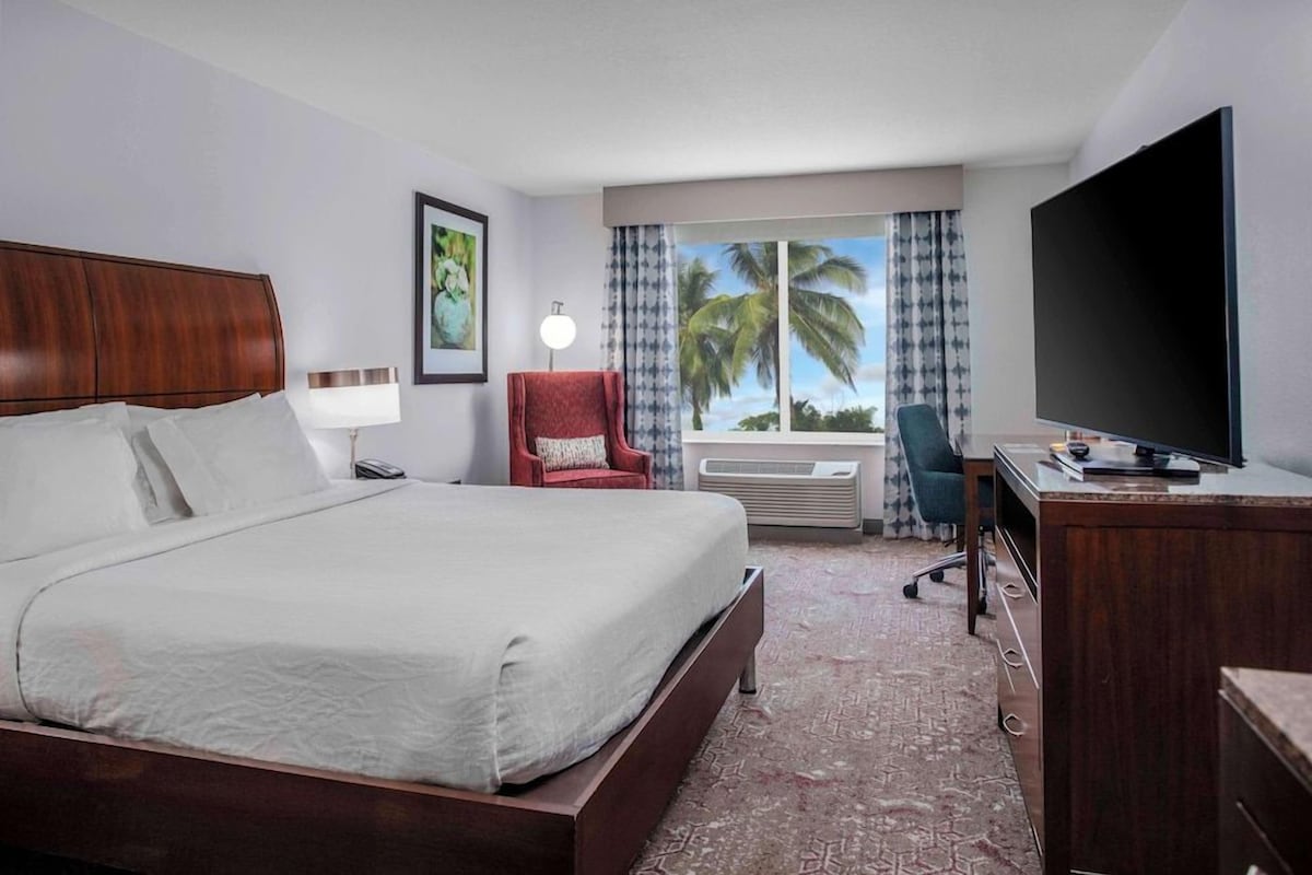 Une chambre d’hôtel avec un grand lit, un bureau doté d’un téléviseur à écran plat, une chaise, une lampe et une fenêtre donnant sur des palmiers.
