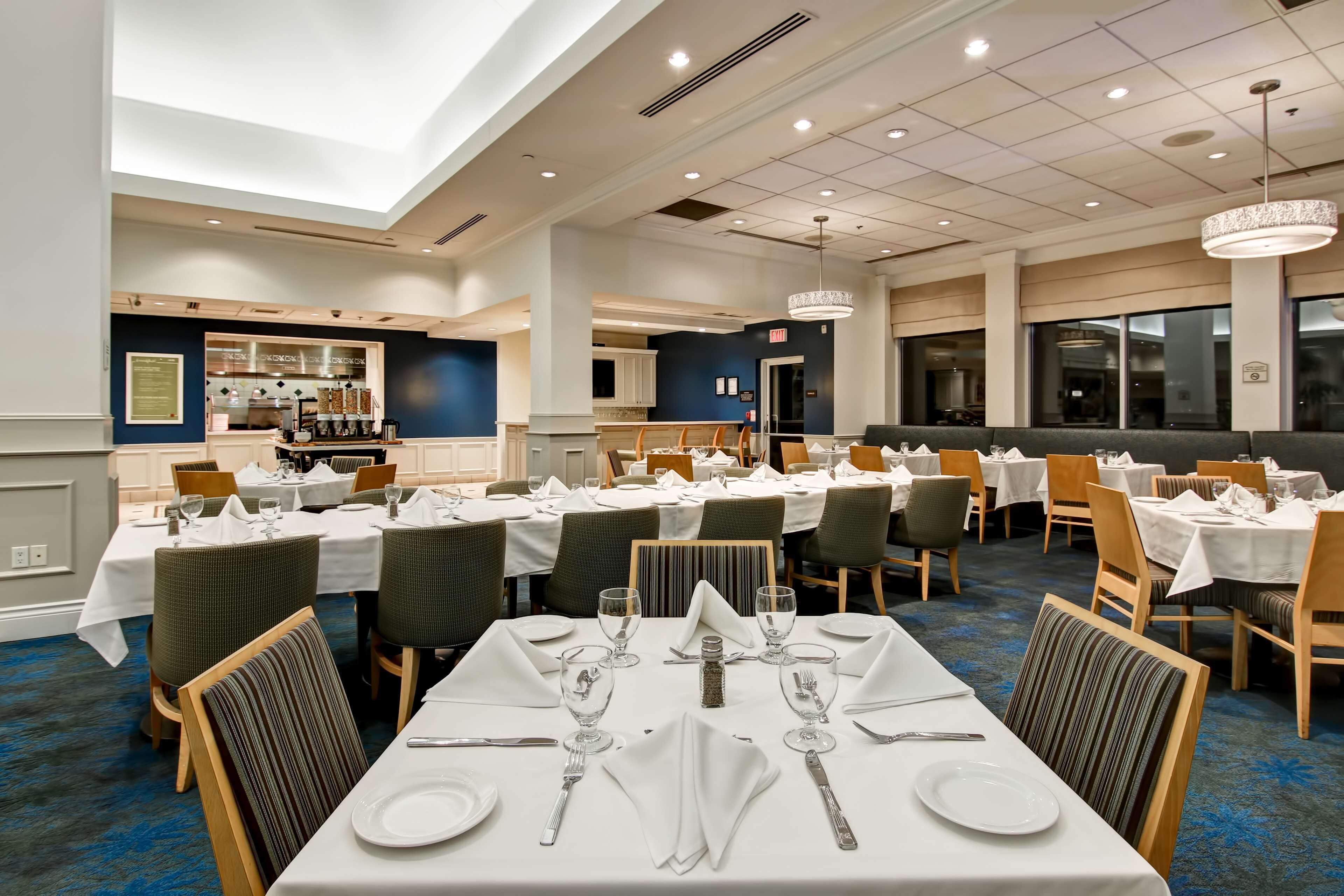 A well-lit dining area with multiple tables set for service, a kitchen in the background, and a high ceiling with recessed lighting.