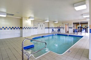 An indoor swimming pool with tiled walls, a blue tiled border, and a pool ladder. There are chairs and a door leading to another room. - Hilton Garden Inn Toronto - Mississauga (Mississauga)