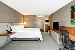 A hotel room with a bed, bedside table, lamp, TV, desk, chair, and a kitchenette. - Hilton Garden Inn Toronto - Mississauga (Mississauga)