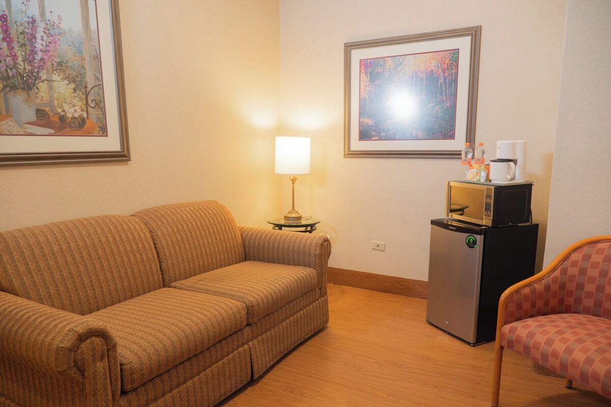 A furnished room with a sofa, a chair, a small table with a lamp, a refrigerator, and two framed pictures on the wall.