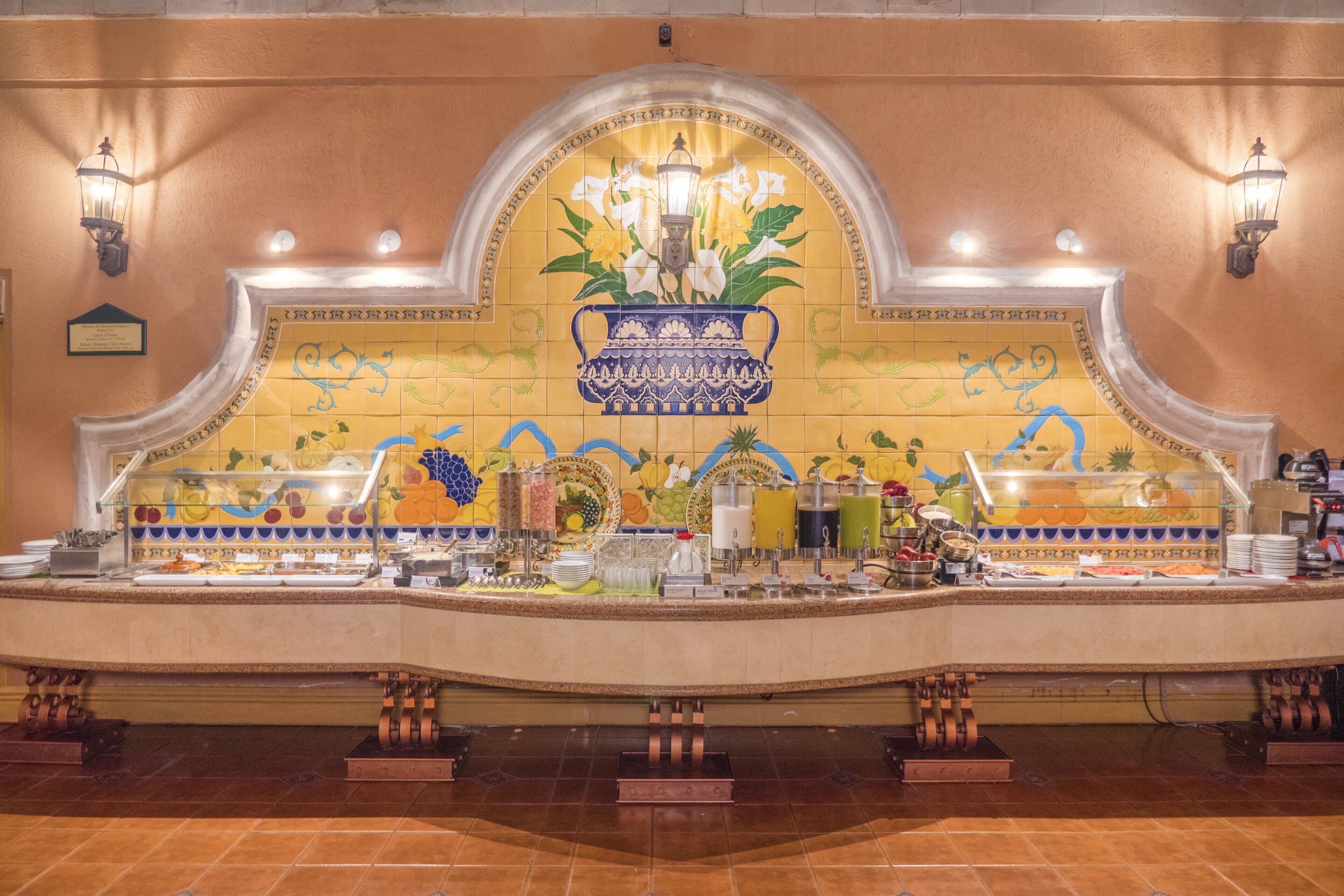 Un área de bufé con salpicadero de azulejos con motivos de frutas y flores, un jarrón decorativo en el centro y varios dispensadores de comida y bebida.