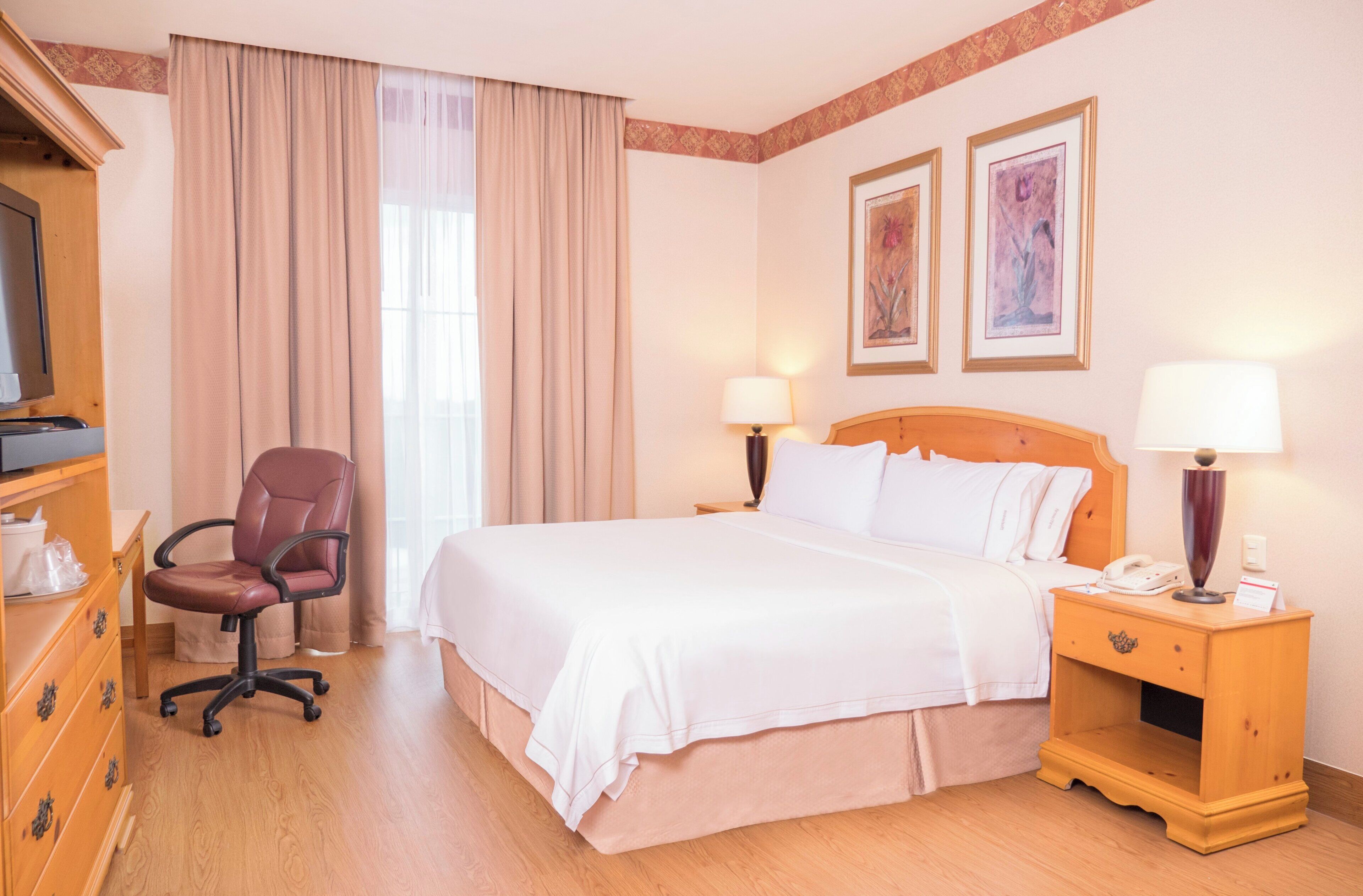 Quarto de hotel com uma cama, escrivaninha, cadeira, abajur e duas imagens emolduradas na parede.