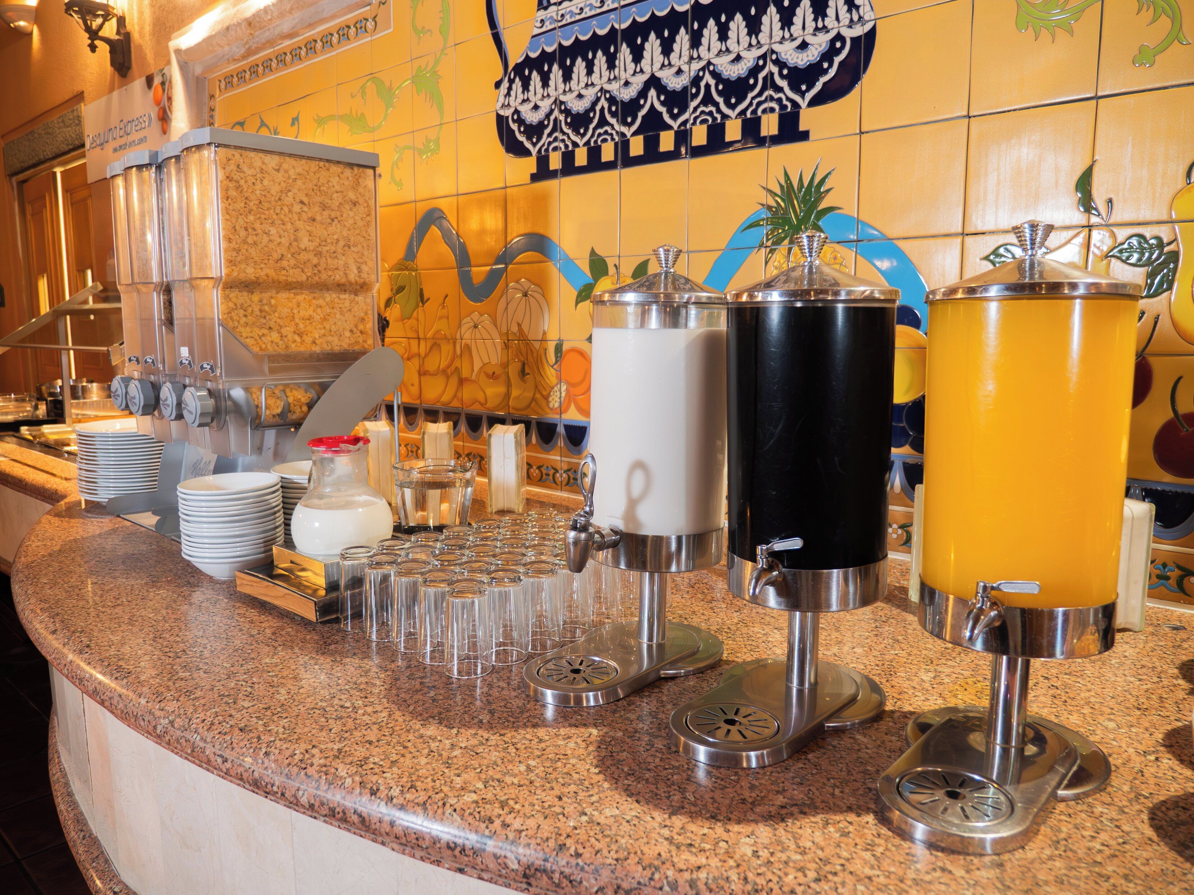 Una barra de buffet con distintos dispensadores de comida y bebidas, una pila de platos y una pared decorativa con estampado floral.